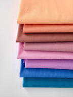 A vertical stack of eight folded fabric pieces showing a color gradient. The colors start at the top with light peach, transitioning through coral, terracotta, and dusty rose, moving into lilac and vibrant purple, and finishing at the bottom with deep royal blue and dark teal. The fabrics are set against a plain white background.