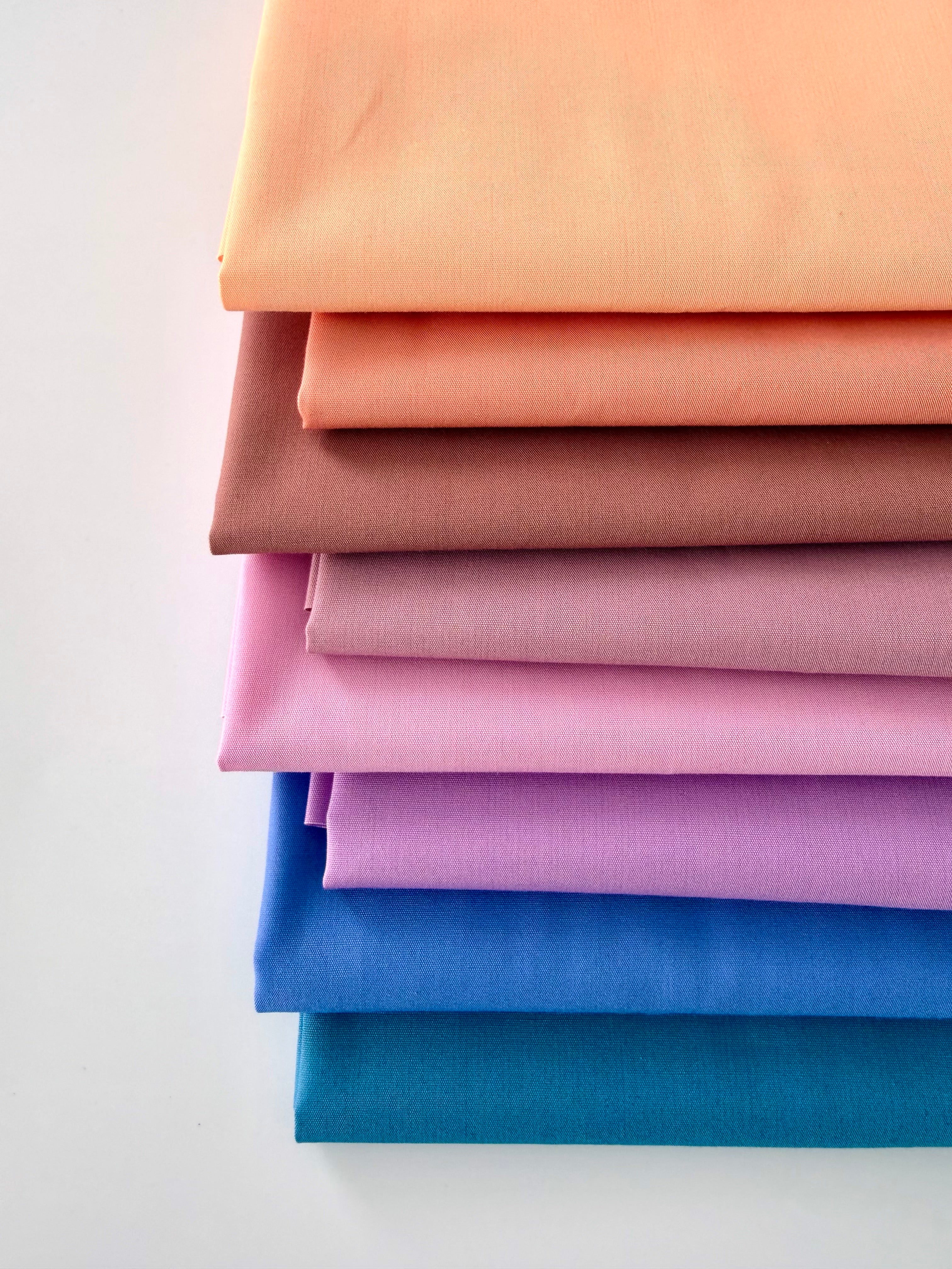A vertical stack of eight folded fabric pieces showing a color gradient. The colors start at the top with light peach, transitioning through coral, terracotta, and dusty rose, moving into lilac and vibrant purple, and finishing at the bottom with deep royal blue and dark teal. The fabrics are set against a plain white background.