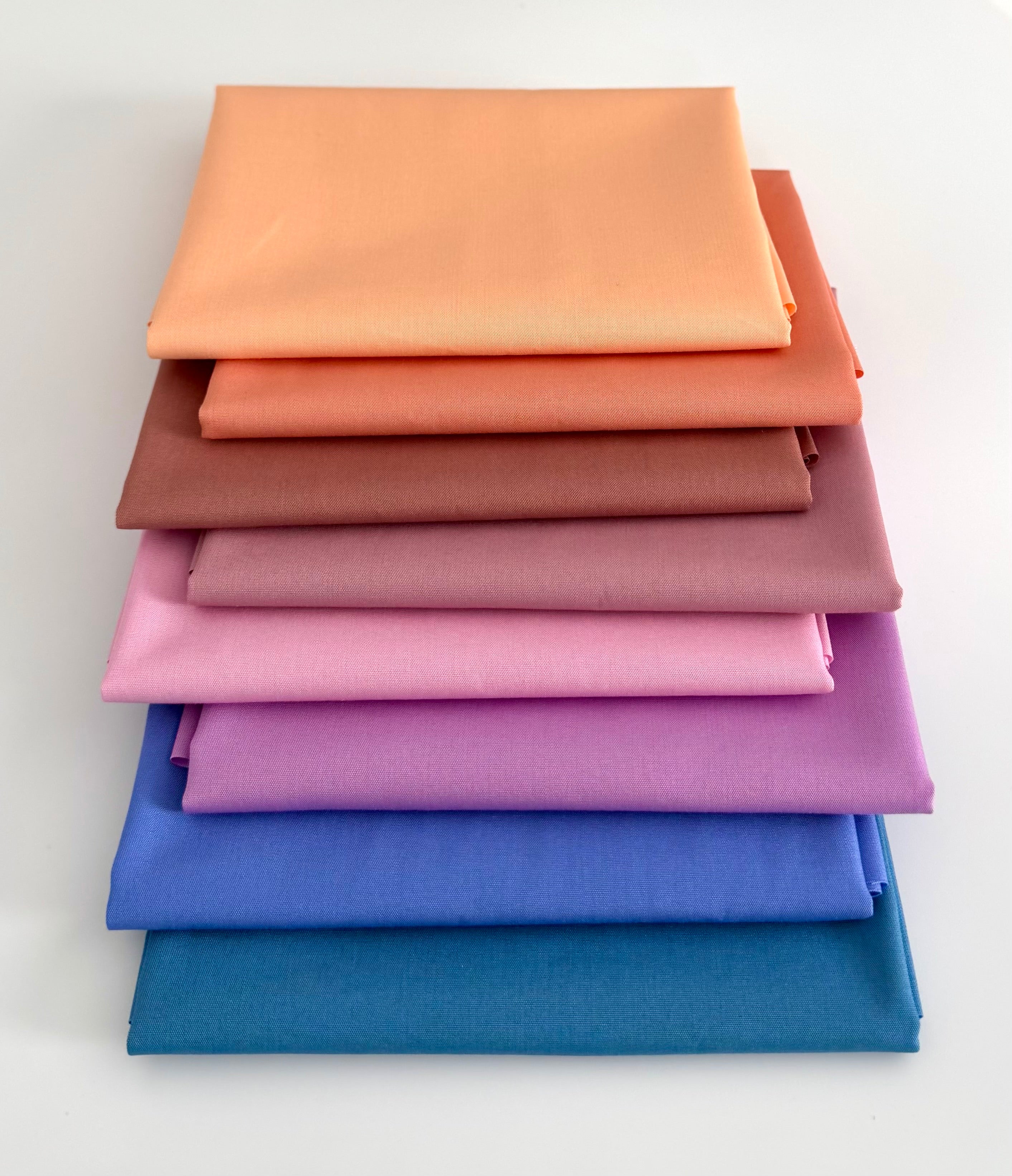 A vertical stack of eight folded fabric pieces showing a color gradient. The colors start at the top with light peach, transitioning through coral, terracotta, and dusty rose, moving into lilac and vibrant purple, and finishing at the bottom with deep royal blue and dark teal. The fabrics are set against a plain white background.