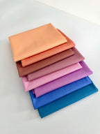 A vertical stack of eight folded fabric pieces showing a color gradient. The colors start at the top with light peach, transitioning through coral, terracotta, and dusty rose, moving into lilac and vibrant purple, and finishing at the bottom with deep royal blue and dark teal. The fabrics are set against a plain white background.