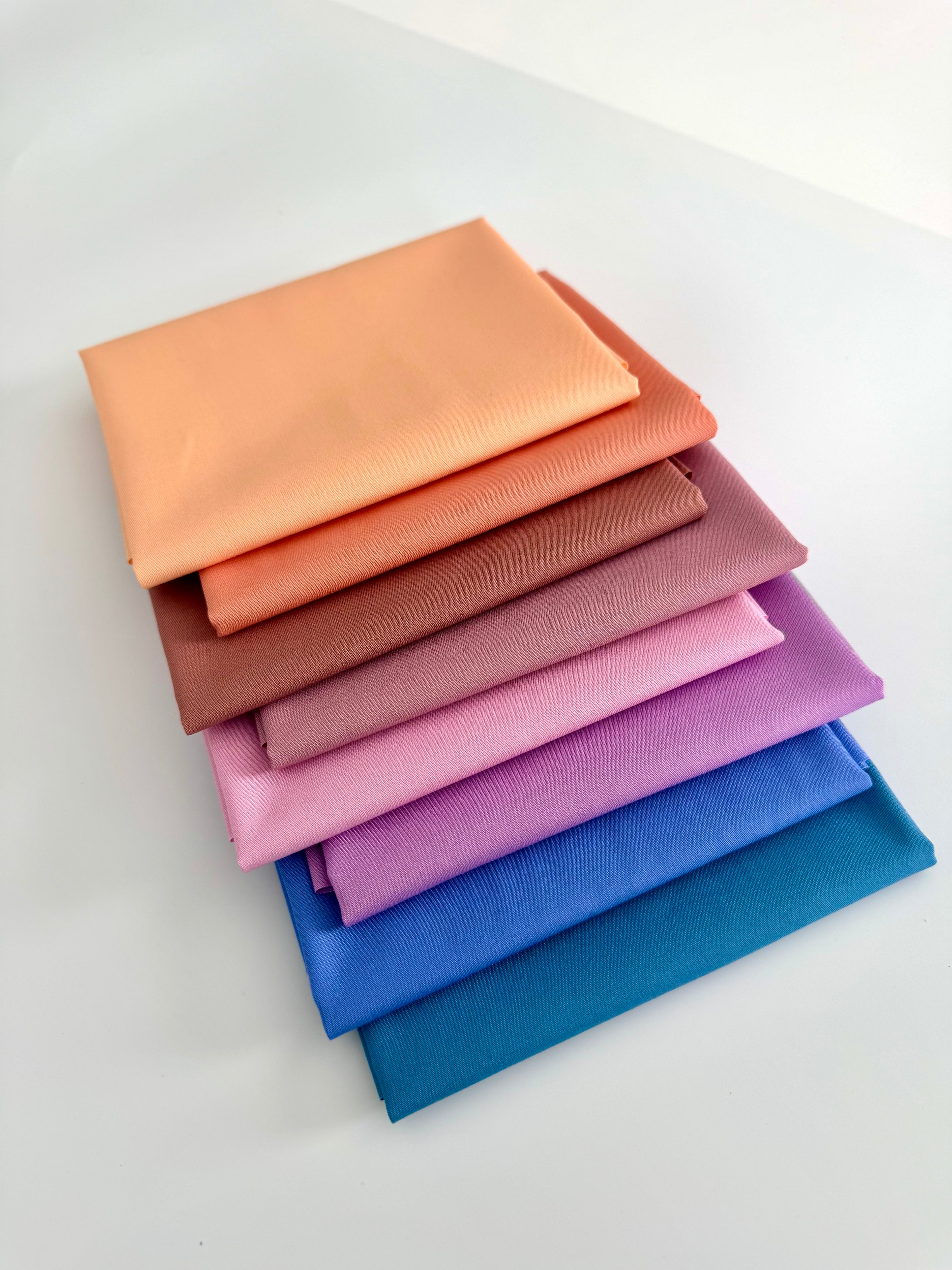 A vertical stack of eight folded fabric pieces showing a color gradient. The colors start at the top with light peach, transitioning through coral, terracotta, and dusty rose, moving into lilac and vibrant purple, and finishing at the bottom with deep royal blue and dark teal. The fabrics are set against a plain white background.
