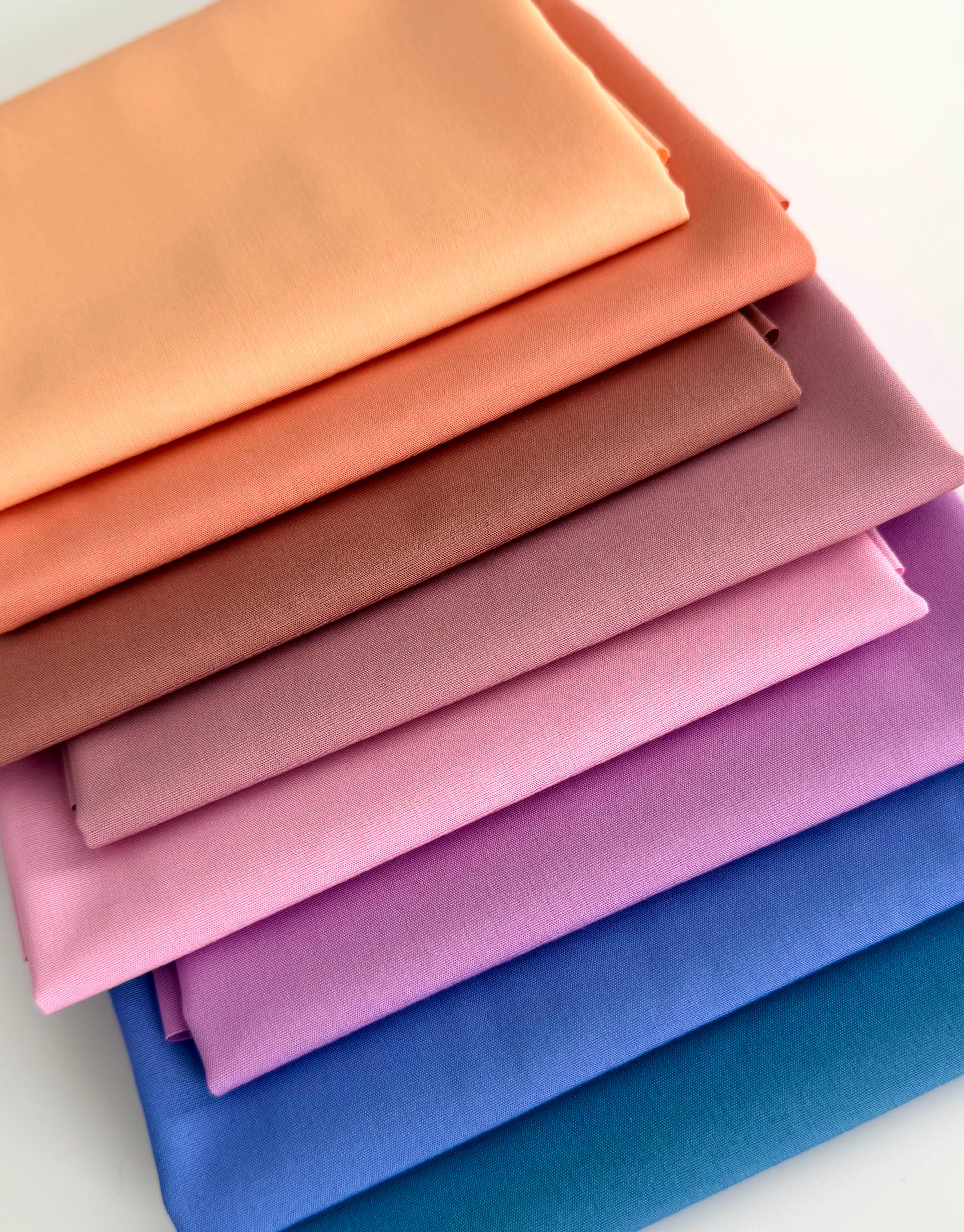 A vertical stack of eight folded fabric pieces showing a color gradient. The colors start at the top with light peach, transitioning through coral, terracotta, and dusty rose, moving into lilac and vibrant purple, and finishing at the bottom with deep royal blue and dark teal. The fabrics are set against a plain white background.