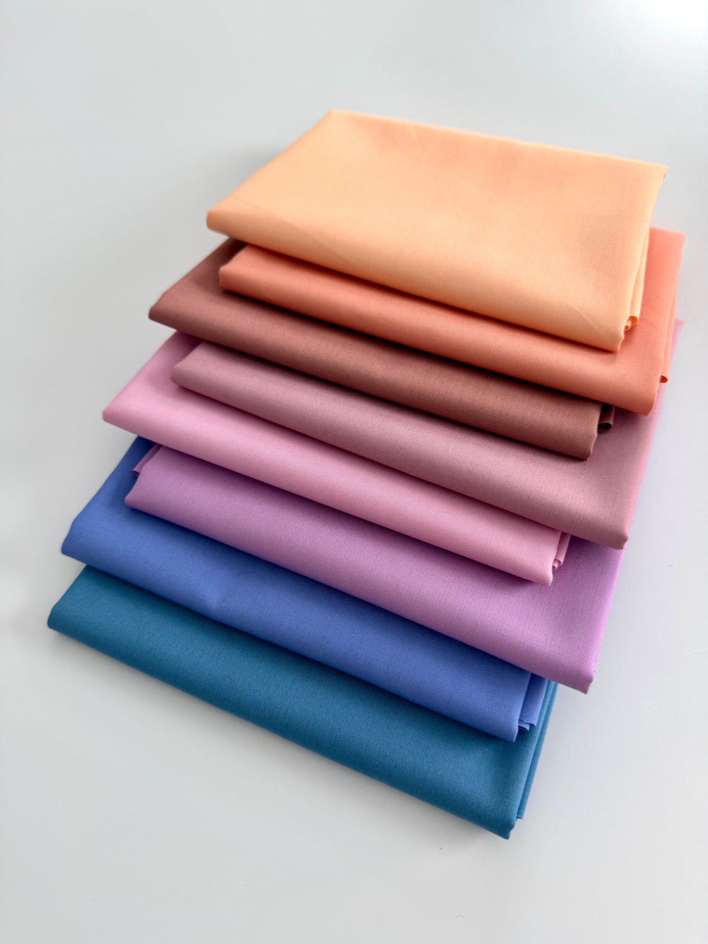 A vertical stack of eight folded fabric pieces showing a color gradient. The colors start at the top with light peach, transitioning through coral, terracotta, and dusty rose, moving into lilac and vibrant purple, and finishing at the bottom with deep royal blue and dark teal. The fabrics are set against a plain white background.