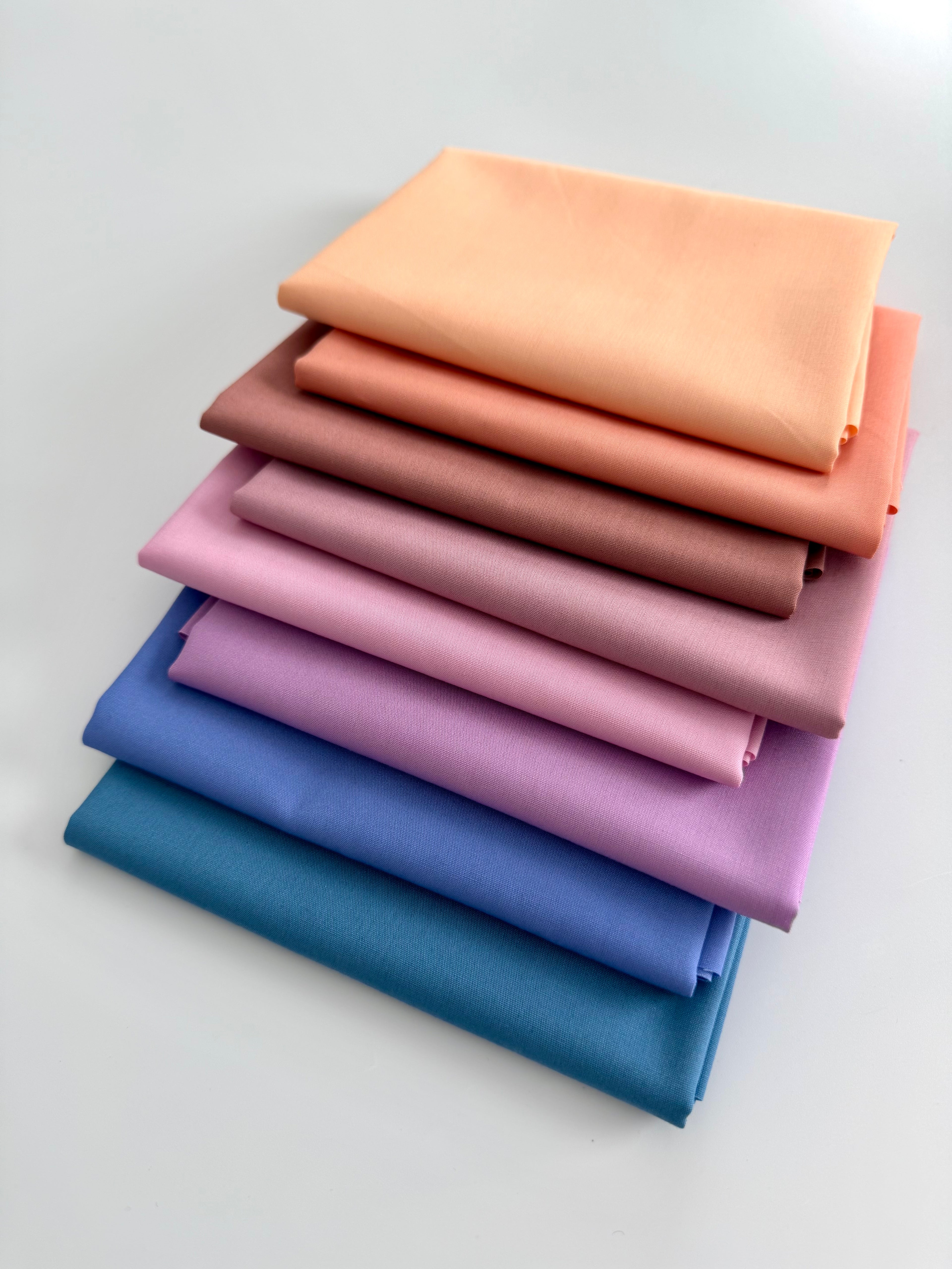 A vertical stack of eight folded fabric pieces showing a color gradient. The colors start at the top with light peach, transitioning through coral, terracotta, and dusty rose, moving into lilac and vibrant purple, and finishing at the bottom with deep royal blue and dark teal. The fabrics are set against a plain white background.