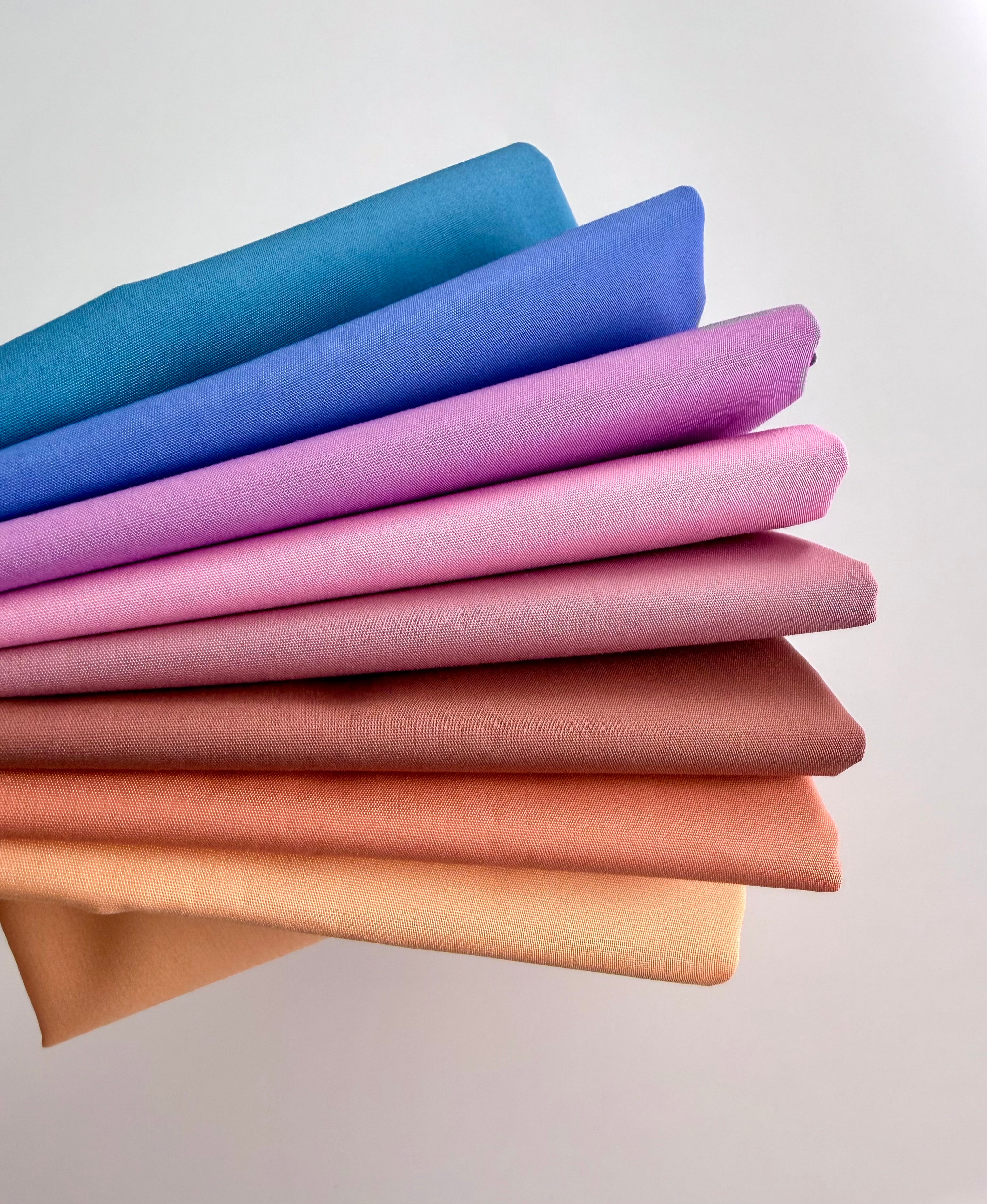 Stack of colorful fabric swatches in blue, purple, pink, brown, and beige on a light gray background.