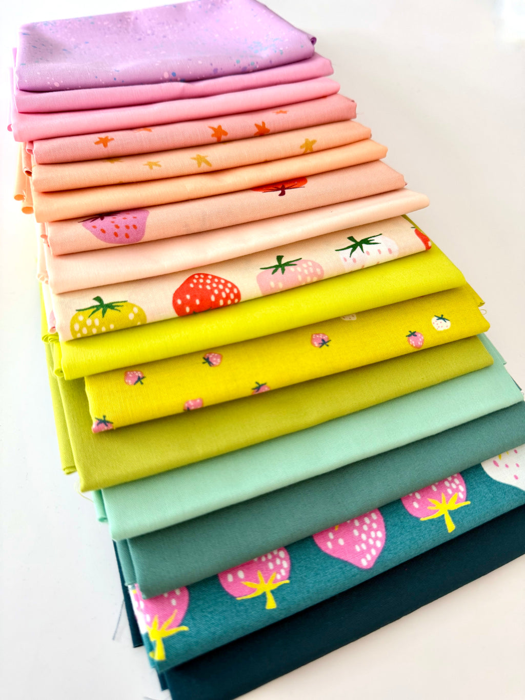 A bright, overhead shot of sixteen overlapping folded fabric pieces. Features a gradient of lavender, bubblegum pink, and peach fabrics with star and strawberry prints, a large-scale strawberry print on cream, transitioning into zesty lime greens, mint, and deep teal fabrics with pink strawberry motifs. The entire collection is displayed against a clean white background.