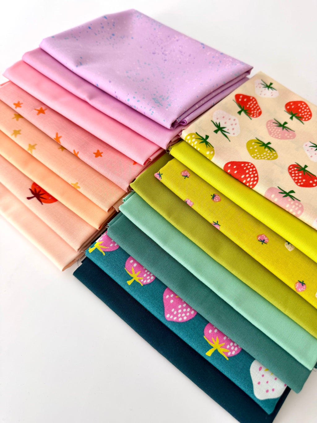A bright, overhead shot of sixteen overlapping folded fabric pieces. Features a gradient of lavender, bubblegum pink, and peach fabrics with star and strawberry prints, a large-scale strawberry print on cream, transitioning into zesty lime greens, mint, and deep teal fabrics with pink strawberry motifs. The entire collection is displayed against a clean white background.