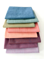 A vertical stack of nine folded Fableism Watercolor Woven fabrics in a "Morning Mist" palette. The colors include a medium dusty blue, followed by sage green, soft ochre, coral, peach, deep plum, muted berry, and finishing with a light lavender and soft coral. The fabrics feature a subtle, yarn-dyed texture with a visible weave, creating a soft, multi-tonal appearance against a clean white background.