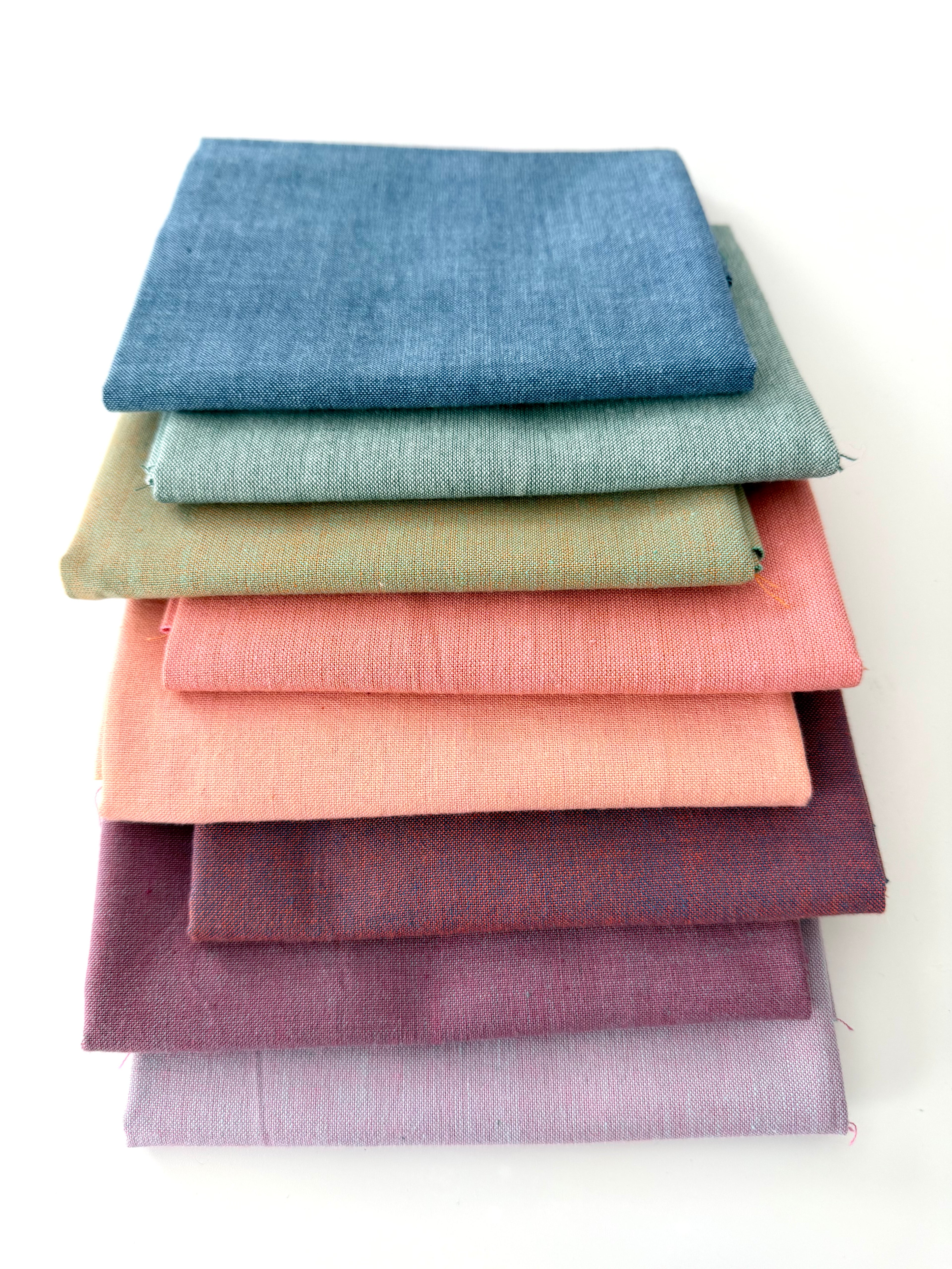 A vertical stack of nine folded Fableism Watercolor Woven fabrics in a "Morning Mist" palette. The colors include a medium dusty blue, followed by sage green, soft ochre, coral, peach, deep plum, muted berry, and finishing with a light lavender and soft coral. The fabrics feature a subtle, yarn-dyed texture with a visible weave, creating a soft, multi-tonal appearance against a clean white background.