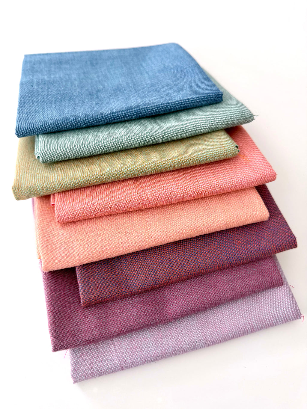 A vertical stack of nine folded Fableism Watercolor Woven fabrics in a "Morning Mist" palette. The colors include a medium dusty blue, followed by sage green, soft ochre, coral, peach, deep plum, muted berry, and finishing with a light lavender and soft coral. The fabrics feature a subtle, yarn-dyed texture with a visible weave, creating a soft, multi-tonal appearance against a clean white background.