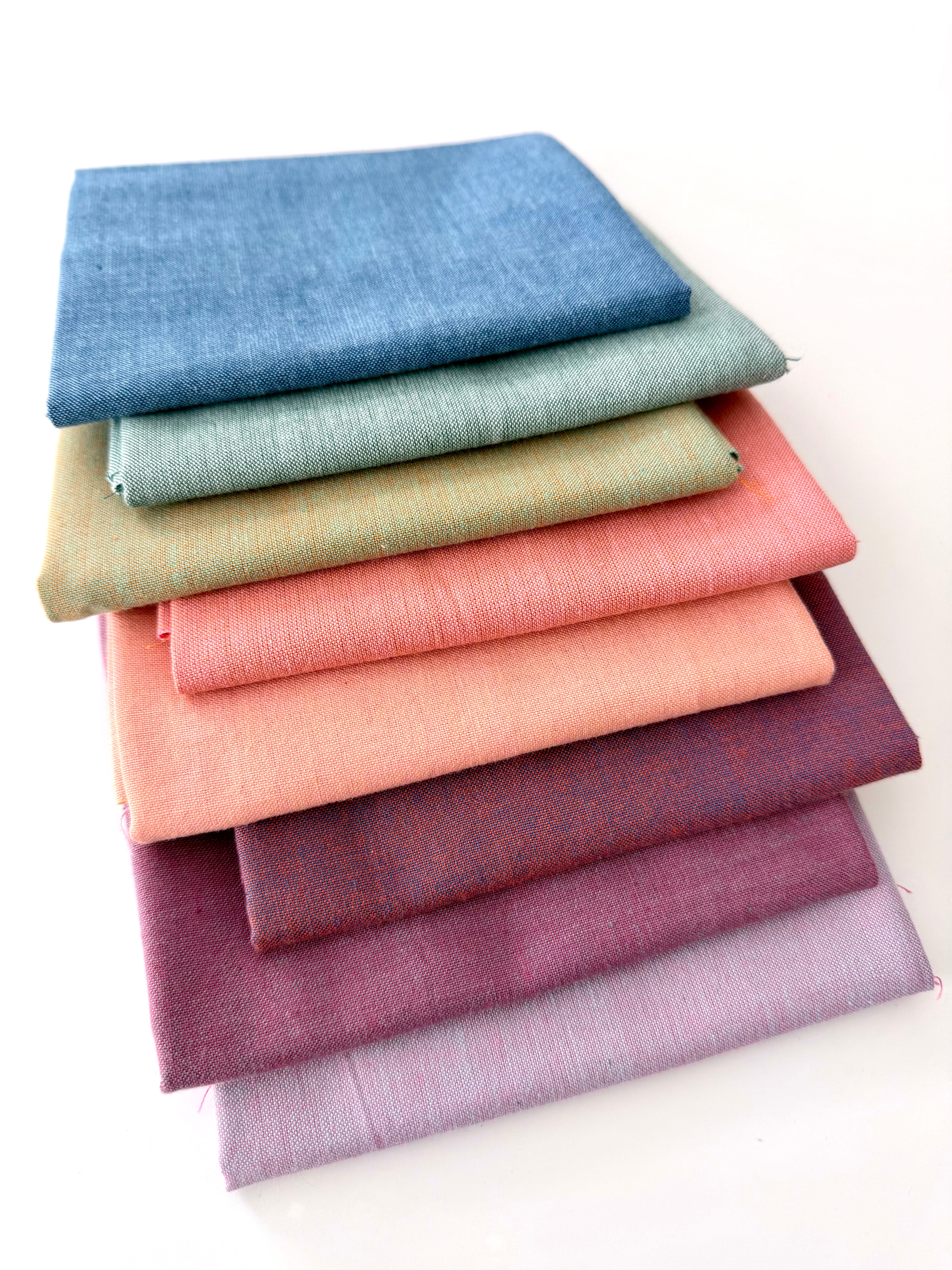 A vertical stack of nine folded Fableism Watercolor Woven fabrics in a "Morning Mist" palette. The colors include a medium dusty blue, followed by sage green, soft ochre, coral, peach, deep plum, muted berry, and finishing with a light lavender and soft coral. The fabrics feature a subtle, yarn-dyed texture with a visible weave, creating a soft, multi-tonal appearance against a clean white background.