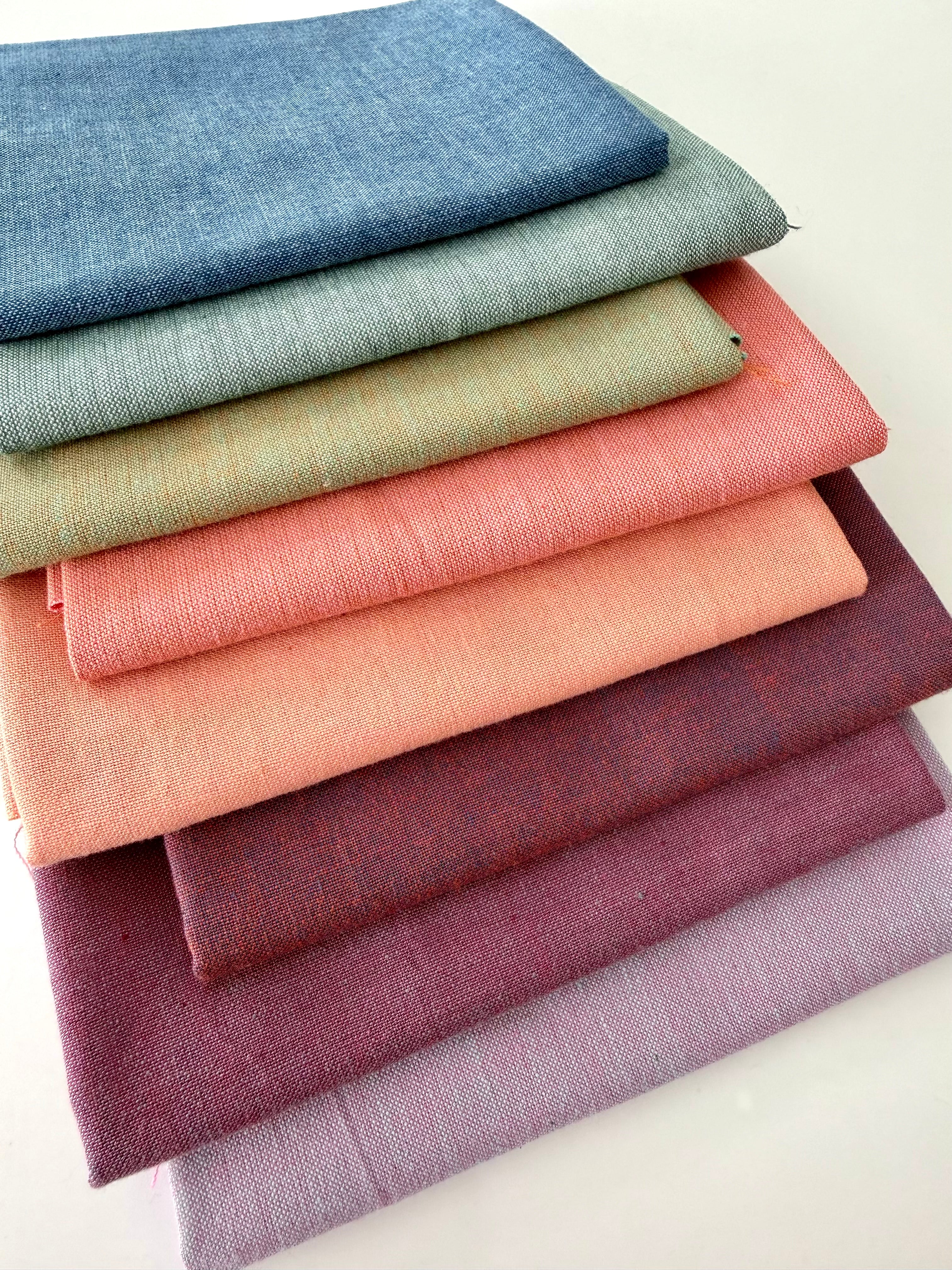 A vertical stack of nine folded Fableism Watercolor Woven fabrics in a "Morning Mist" palette. The colors include a medium dusty blue, followed by sage green, soft ochre, coral, peach, deep plum, muted berry, and finishing with a light lavender and soft coral. The fabrics feature a subtle, yarn-dyed texture with a visible weave, creating a soft, multi-tonal appearance against a clean white background.