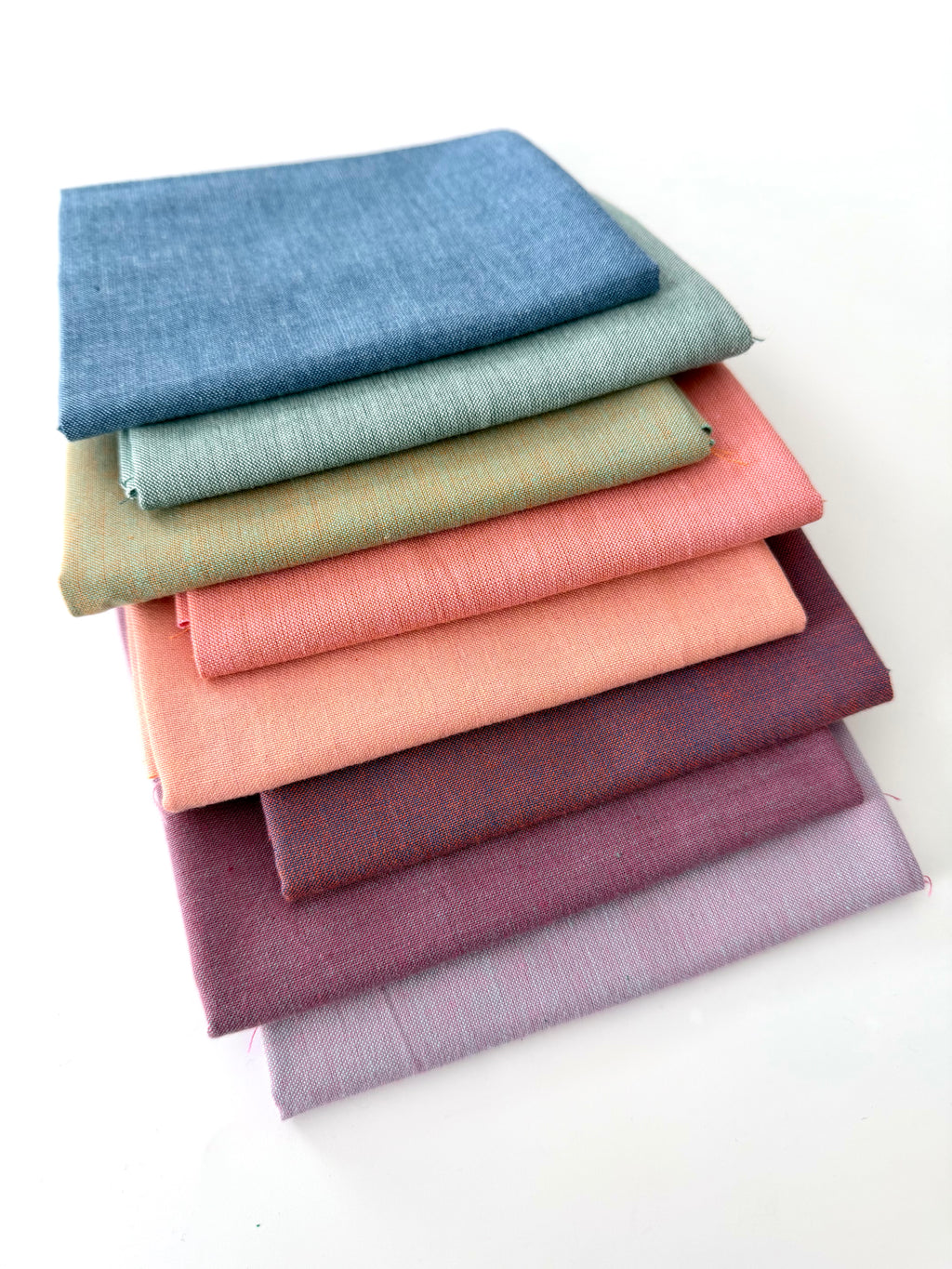 A vertical stack of nine folded Fableism Watercolor Woven fabrics in a "Morning Mist" palette. The colors include a medium dusty blue, followed by sage green, soft ochre, coral, peach, deep plum, muted berry, and finishing with a light lavender and soft coral. The fabrics feature a subtle, yarn-dyed texture with a visible weave, creating a soft, multi-tonal appearance against a clean white background.