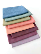A vertical stack of nine folded Fableism Watercolor Woven fabrics in a "Morning Mist" palette. The colors include a medium dusty blue, followed by sage green, soft ochre, coral, peach, deep plum, muted berry, and finishing with a light lavender and soft coral. The fabrics feature a subtle, yarn-dyed texture with a visible weave, creating a soft, multi-tonal appearance against a clean white background.