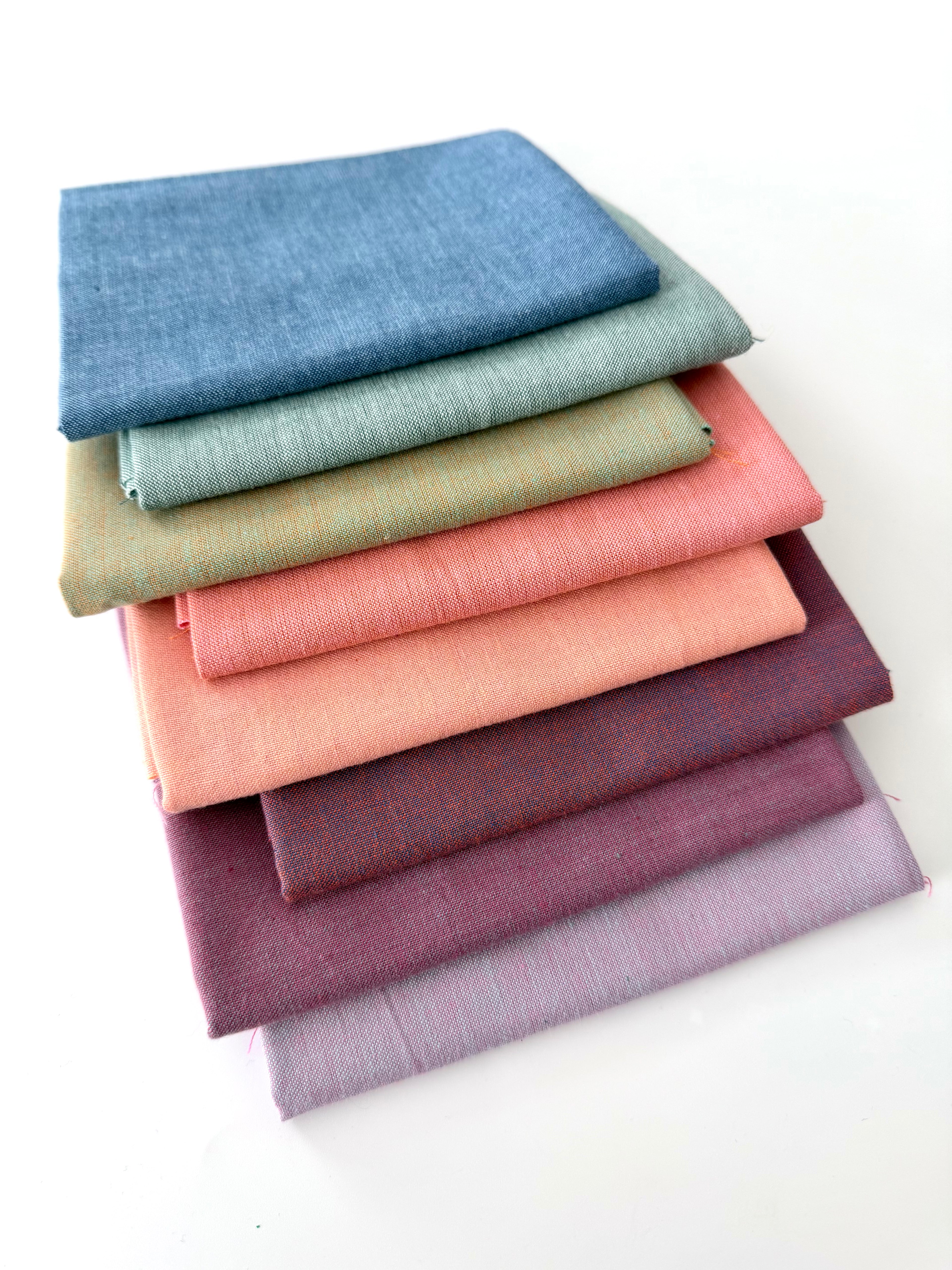 A vertical stack of nine folded Fableism Watercolor Woven fabrics in a "Morning Mist" palette. The colors include a medium dusty blue, followed by sage green, soft ochre, coral, peach, deep plum, muted berry, and finishing with a light lavender and soft coral. The fabrics feature a subtle, yarn-dyed texture with a visible weave, creating a soft, multi-tonal appearance against a clean white background.