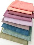 A vertical stack of nine folded Fableism Watercolor Woven fabrics in a "Morning Mist" palette. The colors include a medium dusty blue, followed by sage green, soft ochre, coral, peach, deep plum, muted berry, and finishing with a light lavender and soft coral. The fabrics feature a subtle, yarn-dyed texture with a visible weave, creating a soft, multi-tonal appearance against a clean white background.