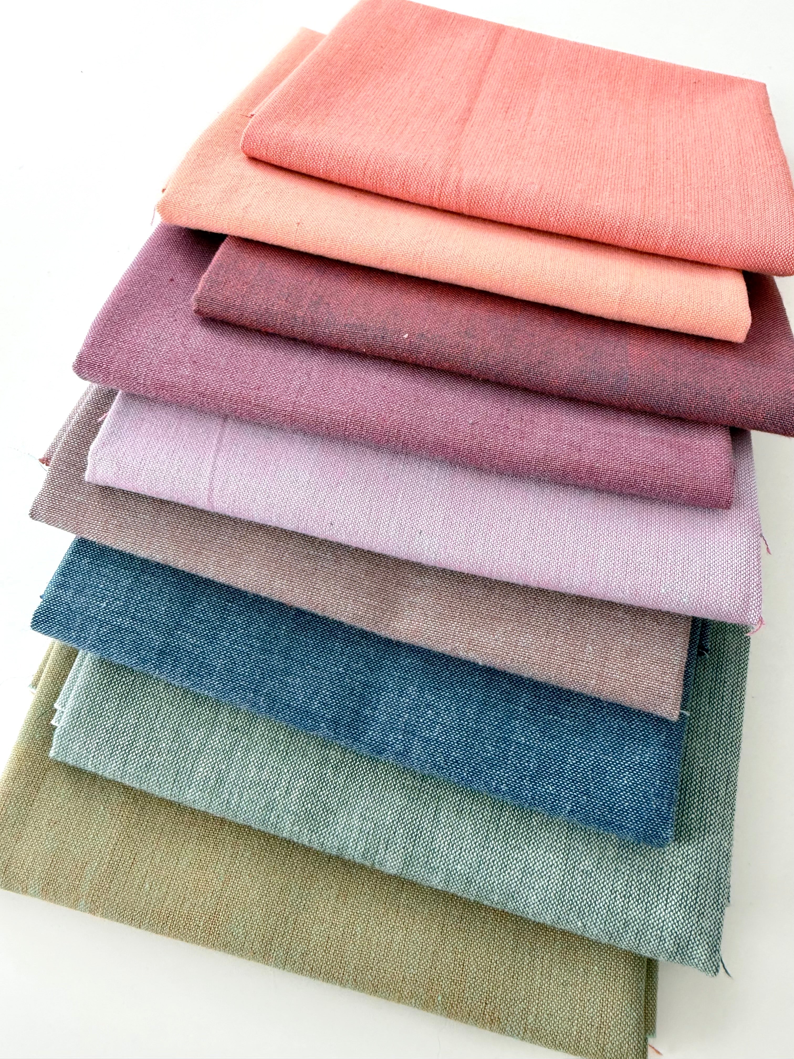 A vertical stack of nine folded Fableism Watercolor Woven fabrics in a "Morning Mist" palette. The colors include a medium dusty blue, followed by sage green, soft ochre, coral, peach, deep plum, muted berry, and finishing with a light lavender and soft coral. The fabrics feature a subtle, yarn-dyed texture with a visible weave, creating a soft, multi-tonal appearance against a clean white background.