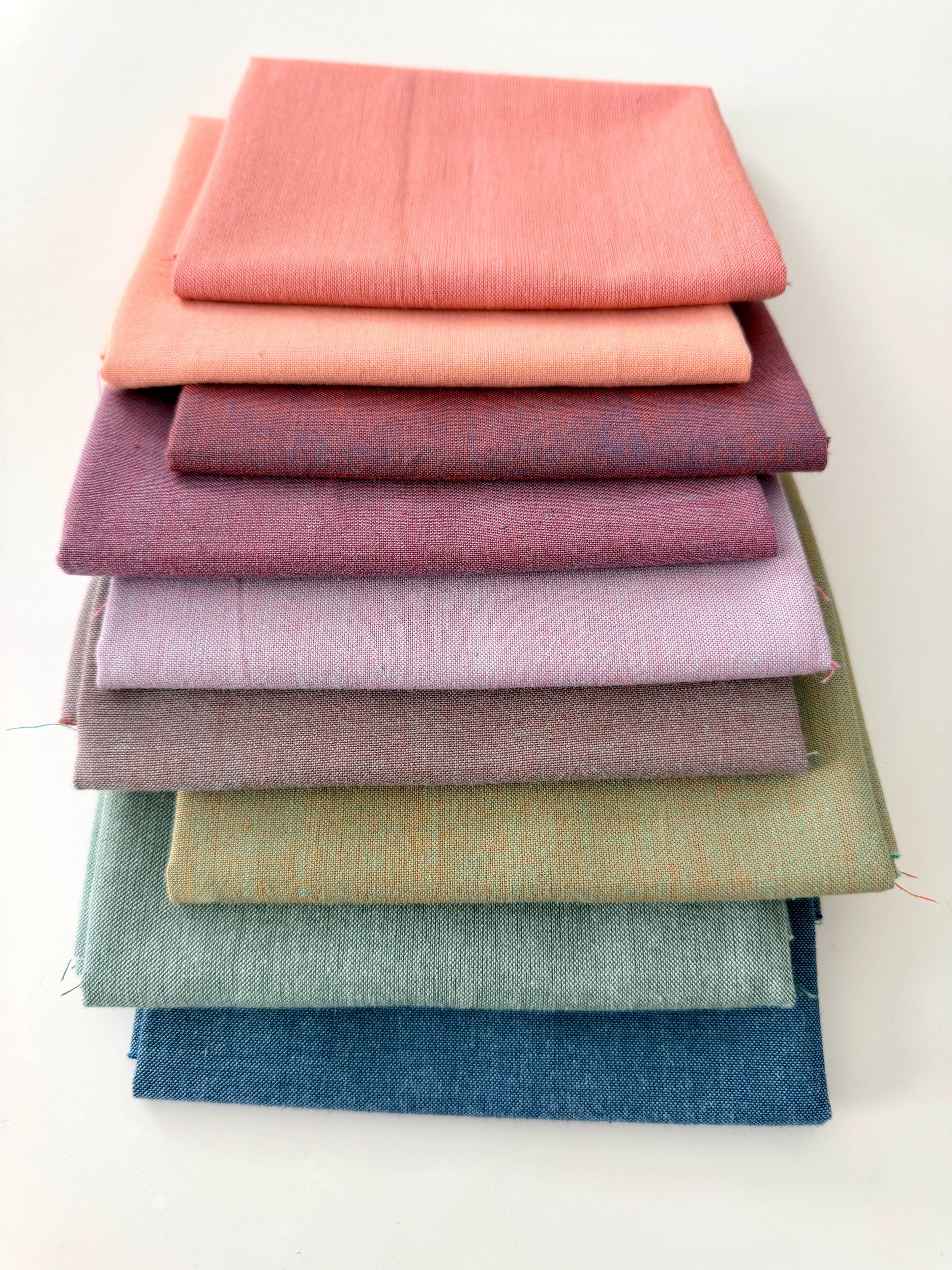 A vertical stack of nine folded Fableism Watercolor Woven fabrics in a "Morning Mist" palette. The colors include a medium dusty blue, followed by sage green, soft ochre, coral, peach, deep plum, muted berry, and finishing with a light lavender and soft coral. The fabrics feature a subtle, yarn-dyed texture with a visible weave, creating a soft, multi-tonal appearance against a clean white background.