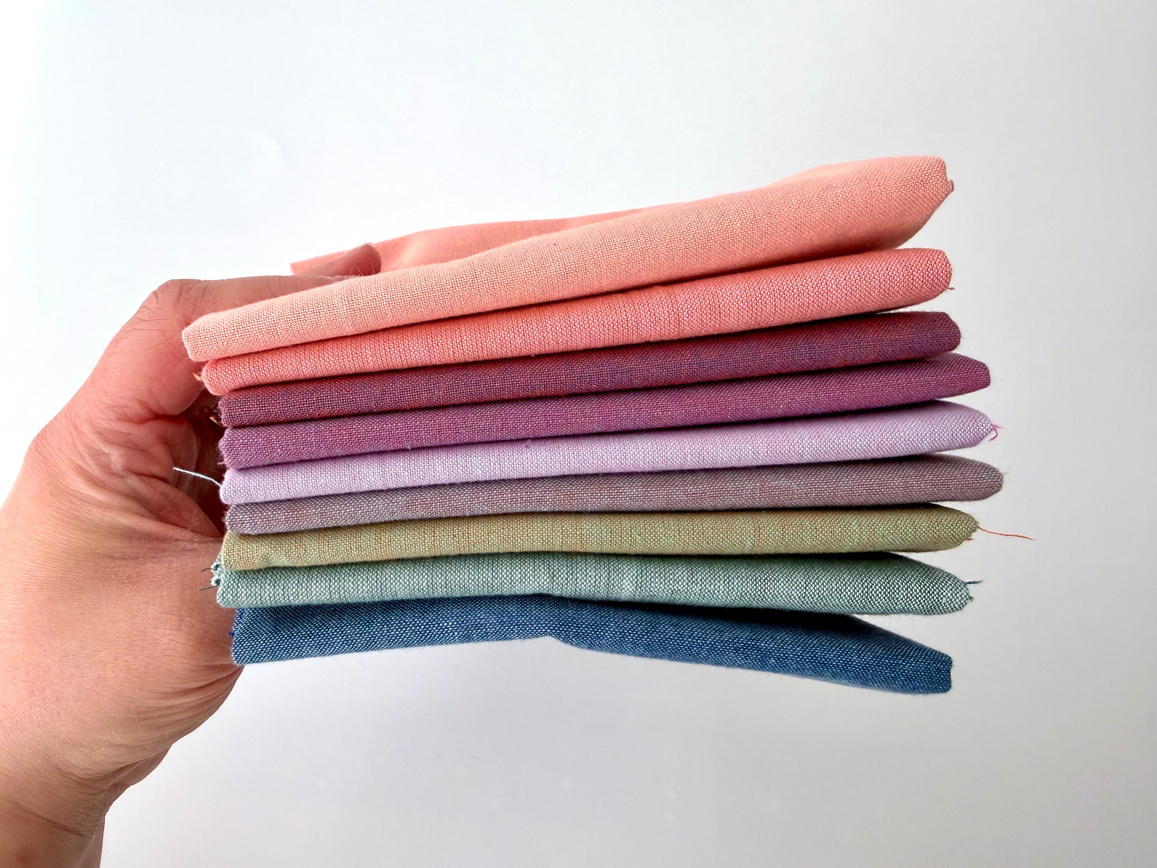 A vertical stack of nine folded Fableism Watercolor Woven fabrics in a "Morning Mist" palette. The colors include a medium dusty blue, followed by sage green, soft ochre, coral, peach, deep plum, muted berry, and finishing with a light lavender and soft coral. The fabrics feature a subtle, yarn-dyed texture with a visible weave, creating a soft, multi-tonal appearance against a clean white background.