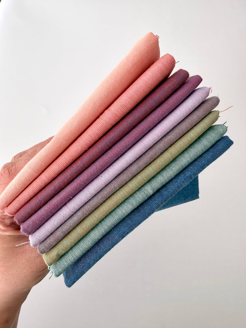 A vertical stack of nine folded Fableism Watercolor Woven fabrics in a "Morning Mist" palette. The colors include a medium dusty blue, followed by sage green, soft ochre, coral, peach, deep plum, muted berry, and finishing with a light lavender and soft coral. The fabrics feature a subtle, yarn-dyed texture with a visible weave, creating a soft, multi-tonal appearance against a clean white background.
