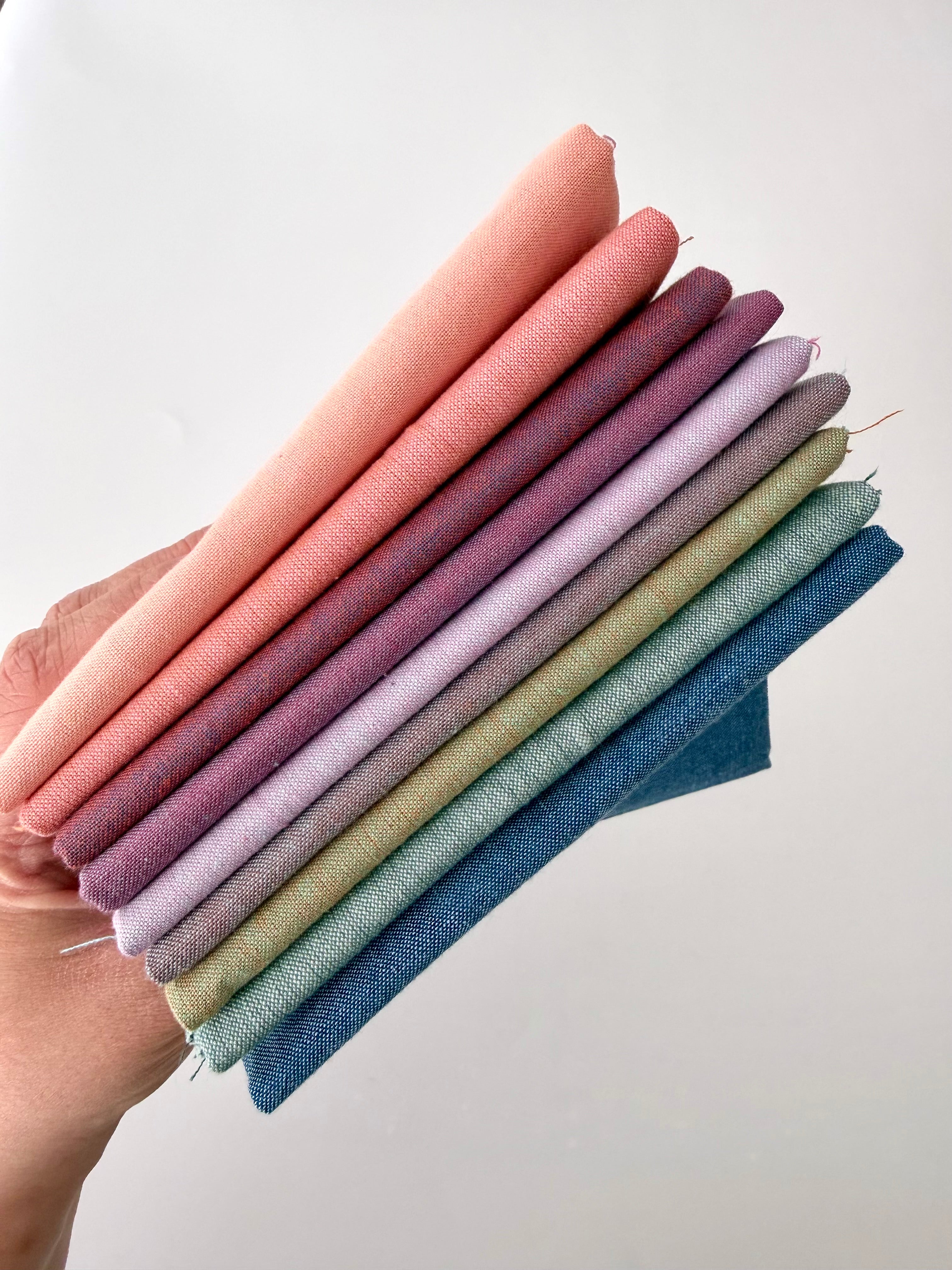 A vertical stack of nine folded Fableism Watercolor Woven fabrics in a "Morning Mist" palette. The colors include a medium dusty blue, followed by sage green, soft ochre, coral, peach, deep plum, muted berry, and finishing with a light lavender and soft coral. The fabrics feature a subtle, yarn-dyed texture with a visible weave, creating a soft, multi-tonal appearance against a clean white background.