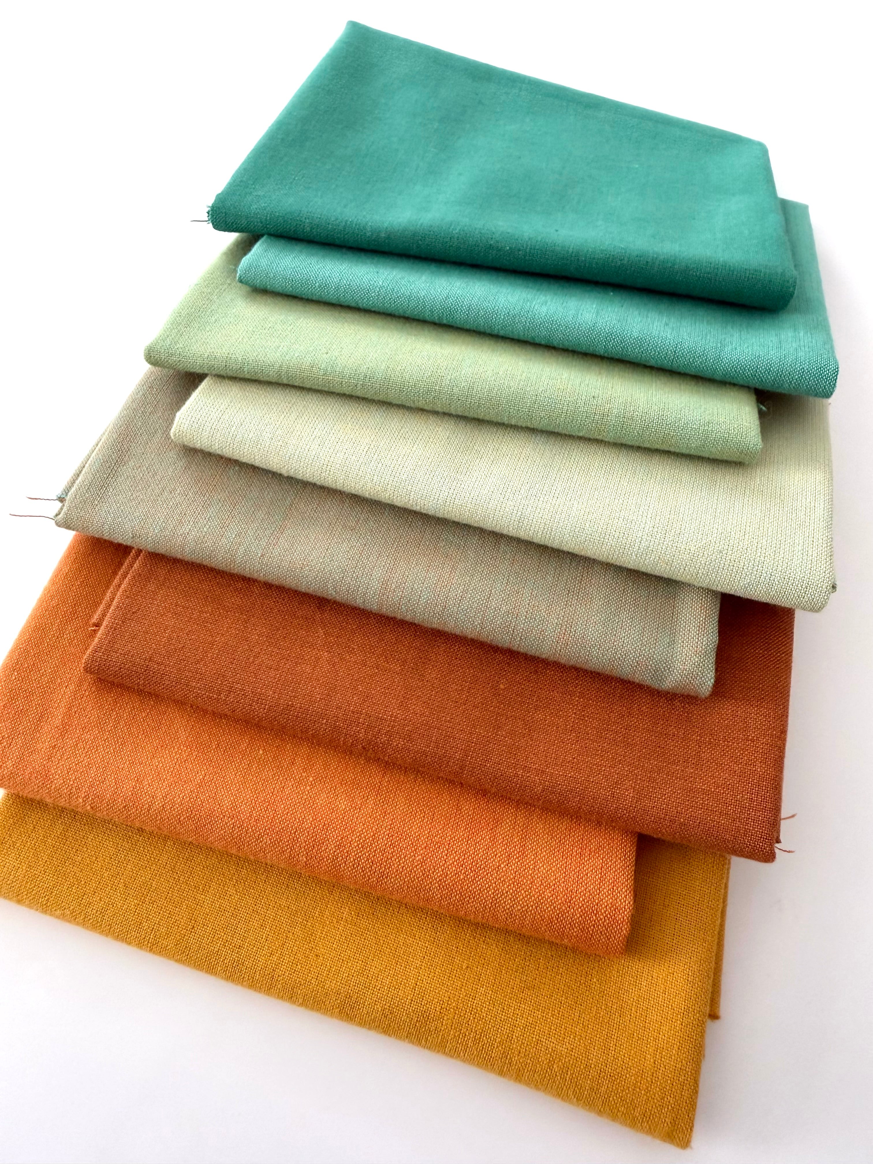 A vertical stack of nine folded fabrics in an earthy gradient, transitioning from teal and sage greens down to terracotta and mustard yellow. The pieces feature a visible yarn-dyed woven texture with subtle grain lines against a white background.