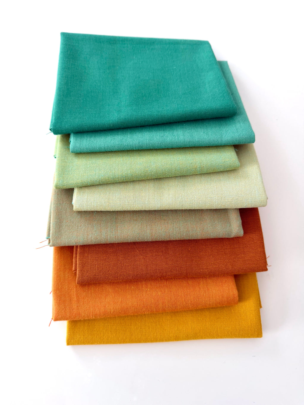 A vertical stack of nine folded fabrics in an earthy gradient, transitioning from teal and sage greens down to terracotta and mustard yellow. The pieces feature a visible yarn-dyed woven texture with subtle grain lines against a white background.