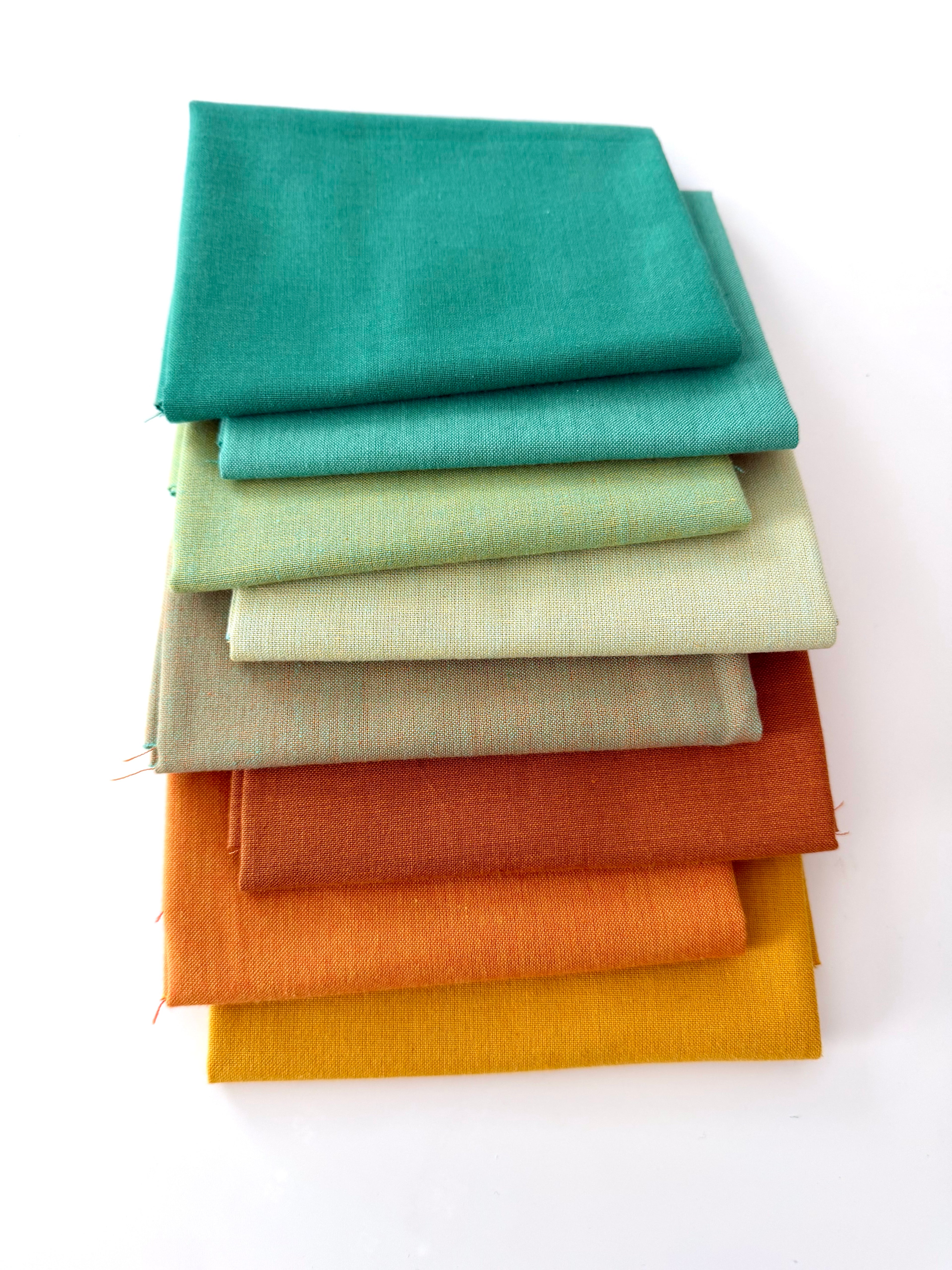 A vertical stack of nine folded fabrics in an earthy gradient, transitioning from teal and sage greens down to terracotta and mustard yellow. The pieces feature a visible yarn-dyed woven texture with subtle grain lines against a white background.