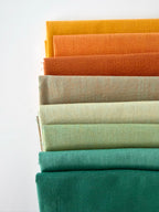 A vertical stack of nine folded fabrics in an earthy gradient, transitioning from teal and sage greens down to terracotta and mustard yellow. The pieces feature a visible yarn-dyed woven texture with subtle grain lines against a white background.