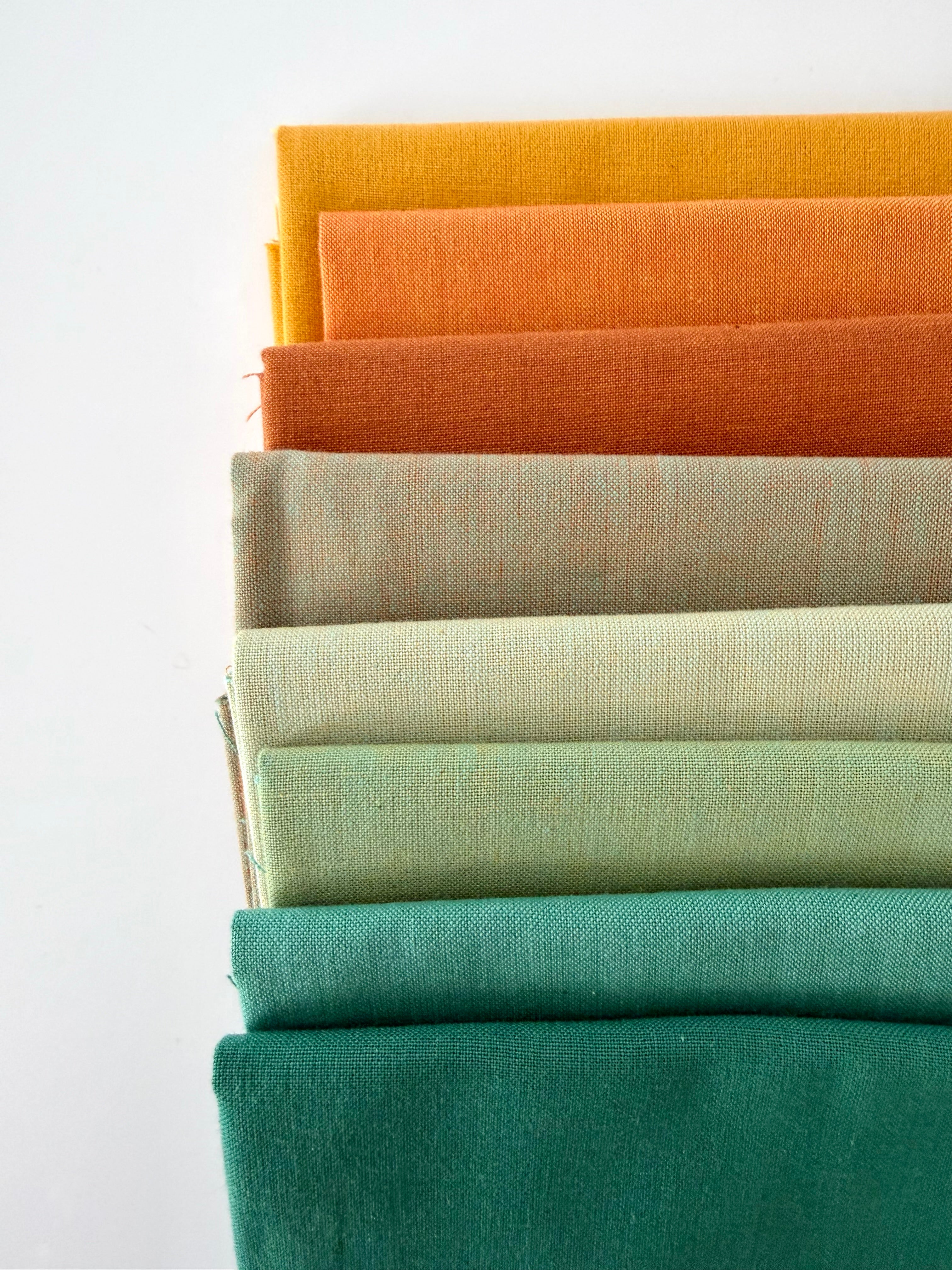 A vertical stack of nine folded fabrics in an earthy gradient, transitioning from teal and sage greens down to terracotta and mustard yellow. The pieces feature a visible yarn-dyed woven texture with subtle grain lines against a white background.