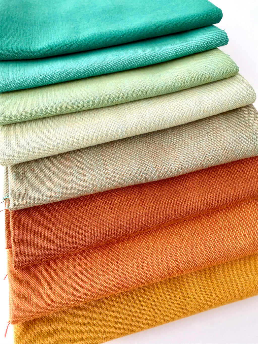 A vertical stack of nine folded fabrics in an earthy gradient, transitioning from teal and sage greens down to terracotta and mustard yellow. The pieces feature a visible yarn-dyed woven texture with subtle grain lines against a white background.