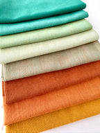 A vertical stack of nine folded fabrics in an earthy gradient, transitioning from teal and sage greens down to terracotta and mustard yellow. The pieces feature a visible yarn-dyed woven texture with subtle grain lines against a white background.
