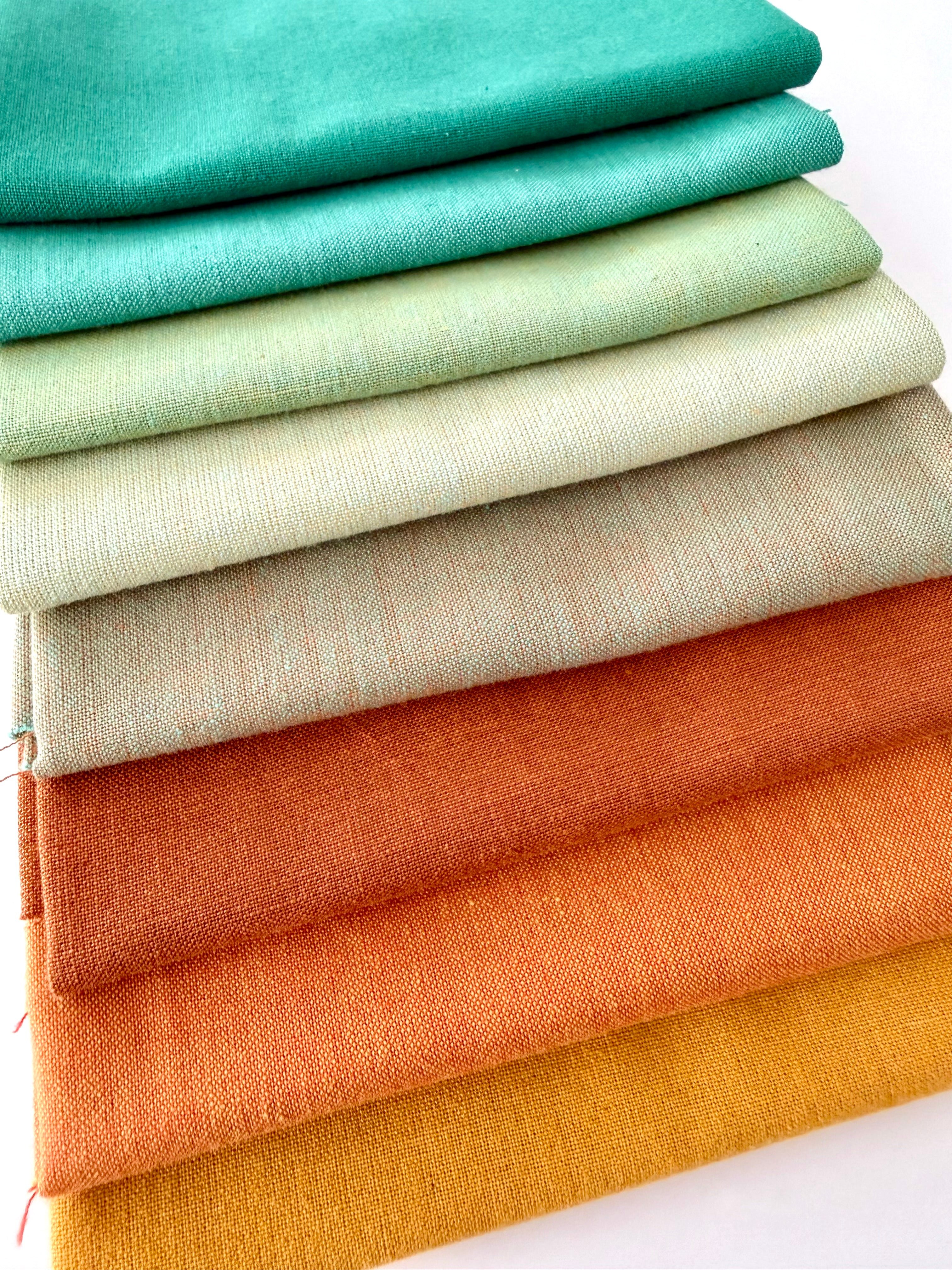 A vertical stack of nine folded fabrics in an earthy gradient, transitioning from teal and sage greens down to terracotta and mustard yellow. The pieces feature a visible yarn-dyed woven texture with subtle grain lines against a white background.