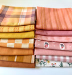 A curated stack of 14 high-quality fabric quarters from the Golden Hour Glow bundle. The mix features rich mustard yellow, deep terracotta rust, soft blush pink, cream, and warm neutral fabrics. The collection showcases a blend of textures and patterns, including yarn-dyed wovens, classic ginghams, windowpane plaids, delicate stripes, and a central floral botanical print, styled flat on a clean white background.
