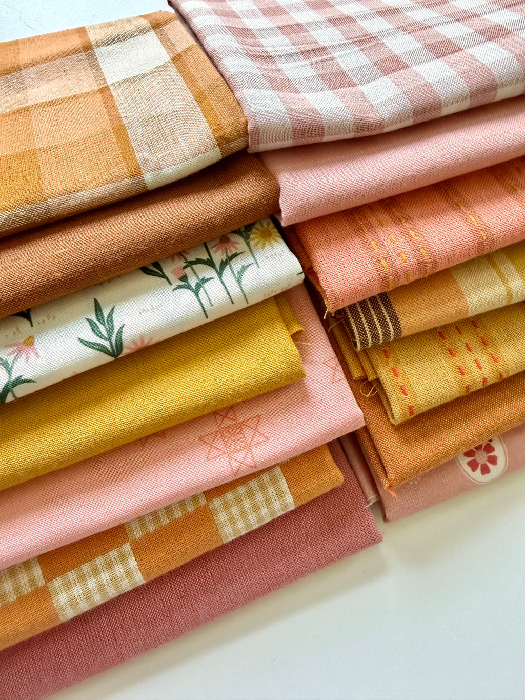 A curated stack of 14 high-quality fabric quarters from the Golden Hour Glow bundle. The mix features rich mustard yellow, deep terracotta rust, soft blush pink, cream, and warm neutral fabrics. The collection showcases a blend of textures and patterns, including yarn-dyed wovens, classic ginghams, windowpane plaids, delicate stripes, and a central floral botanical print, styled flat on a clean white background.