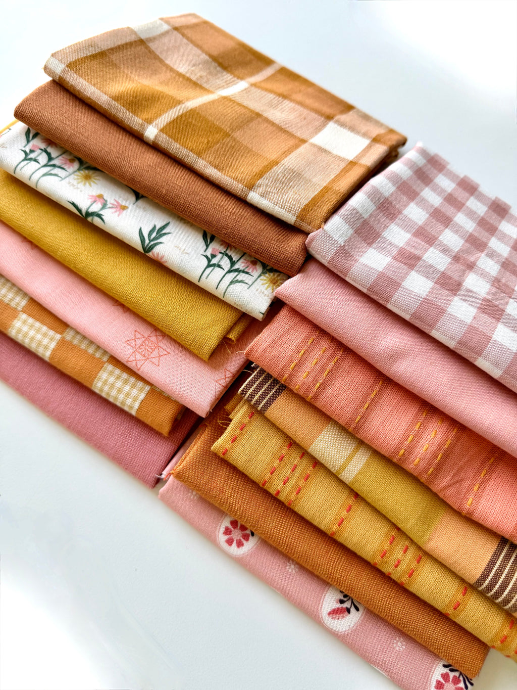 A curated stack of 14 high-quality fabric quarters from the Golden Hour Glow bundle. The mix features rich mustard yellow, deep terracotta rust, soft blush pink, cream, and warm neutral fabrics. The collection showcases a blend of textures and patterns, including yarn-dyed wovens, classic ginghams, windowpane plaids, delicate stripes, and a central floral botanical print, styled flat on a clean white background.