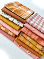 A curated stack of 14 high-quality fabric quarters from the Golden Hour Glow bundle. The mix features rich mustard yellow, deep terracotta rust, soft blush pink, cream, and warm neutral fabrics. The collection showcases a blend of textures and patterns, including yarn-dyed wovens, classic ginghams, windowpane plaids, delicate stripes, and a central floral botanical print, styled flat on a clean white background.