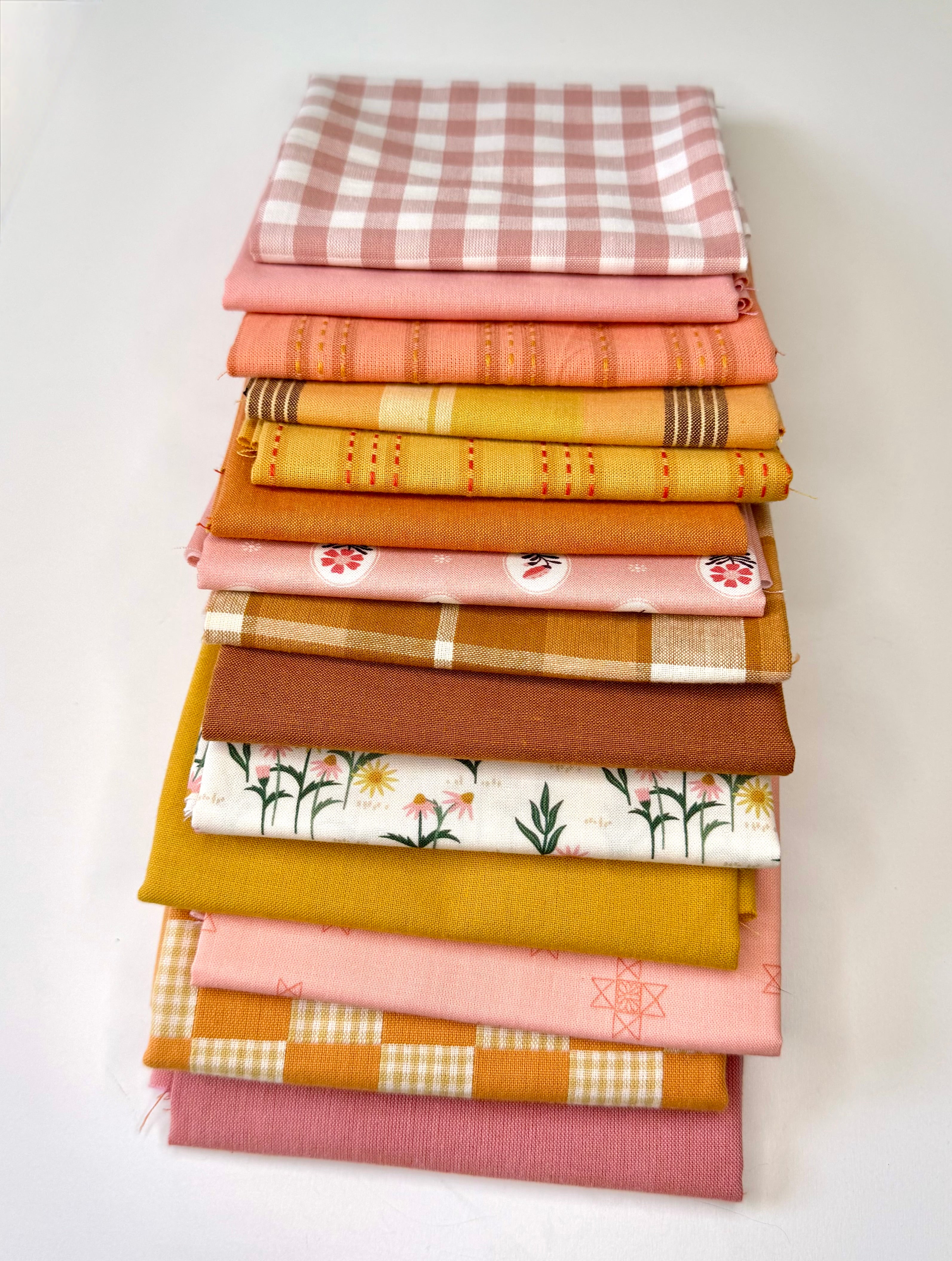 A curated stack of 14 high-quality fabric quarters from the Golden Hour Glow bundle. The mix features rich mustard yellow, deep terracotta rust, soft blush pink, cream, and warm neutral fabrics. The collection showcases a blend of textures and patterns, including yarn-dyed wovens, classic ginghams, windowpane plaids, delicate stripes, and a central floral botanical print, styled flat on a clean white background.