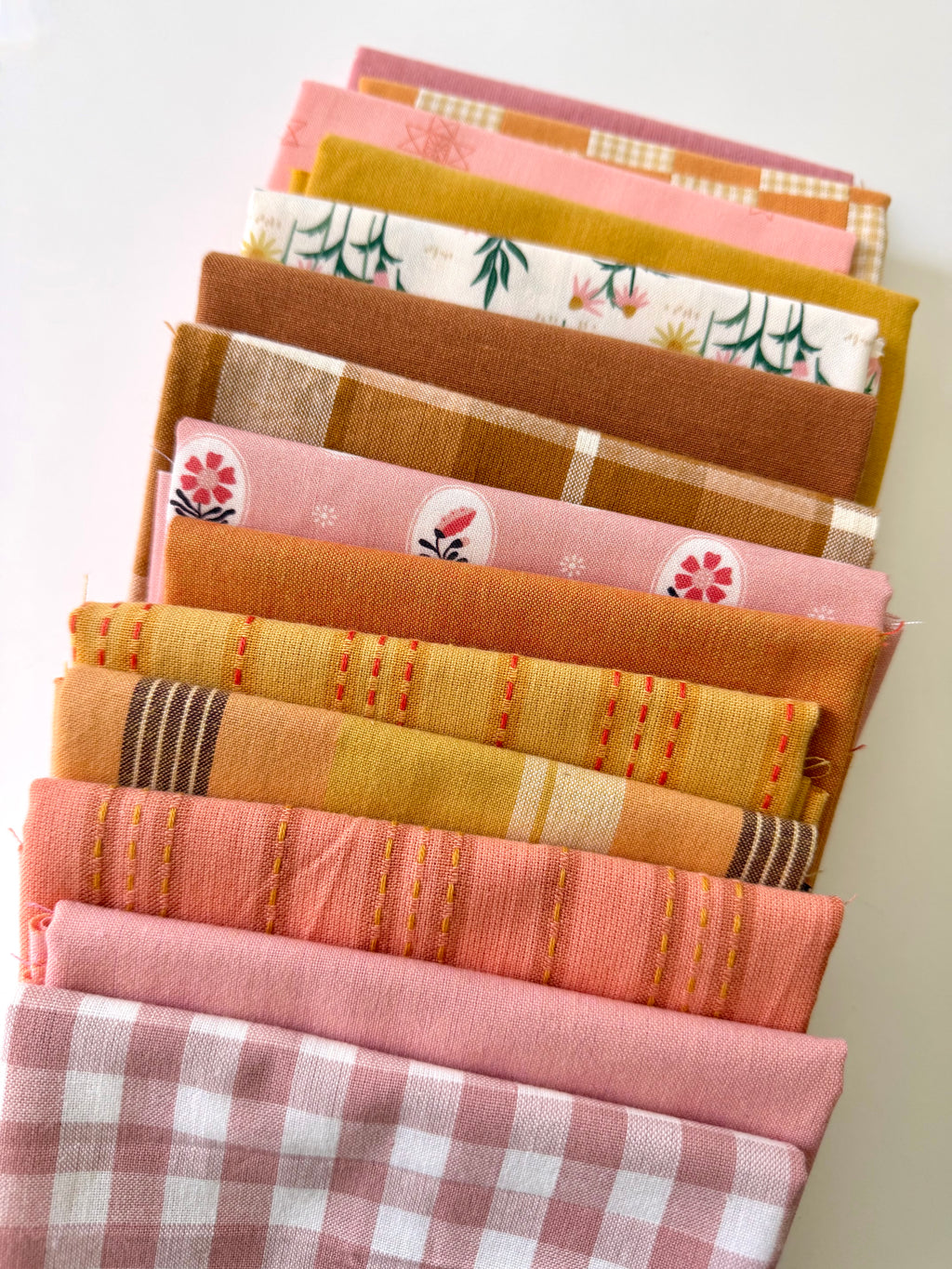 A curated stack of 14 high-quality fabric quarters from the Golden Hour Glow bundle. The mix features rich mustard yellow, deep terracotta rust, soft blush pink, cream, and warm neutral fabrics. The collection showcases a blend of textures and patterns, including yarn-dyed wovens, classic ginghams, windowpane plaids, delicate stripes, and a central floral botanical print, styled flat on a clean white background.