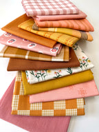 A curated stack of 14 high-quality fabric quarters from the Golden Hour Glow bundle. The mix features rich mustard yellow, deep terracotta rust, soft blush pink, cream, and warm neutral fabrics. The collection showcases a blend of textures and patterns, including yarn-dyed wovens, classic ginghams, windowpane plaids, delicate stripes, and a central floral botanical print, styled flat on a clean white background.