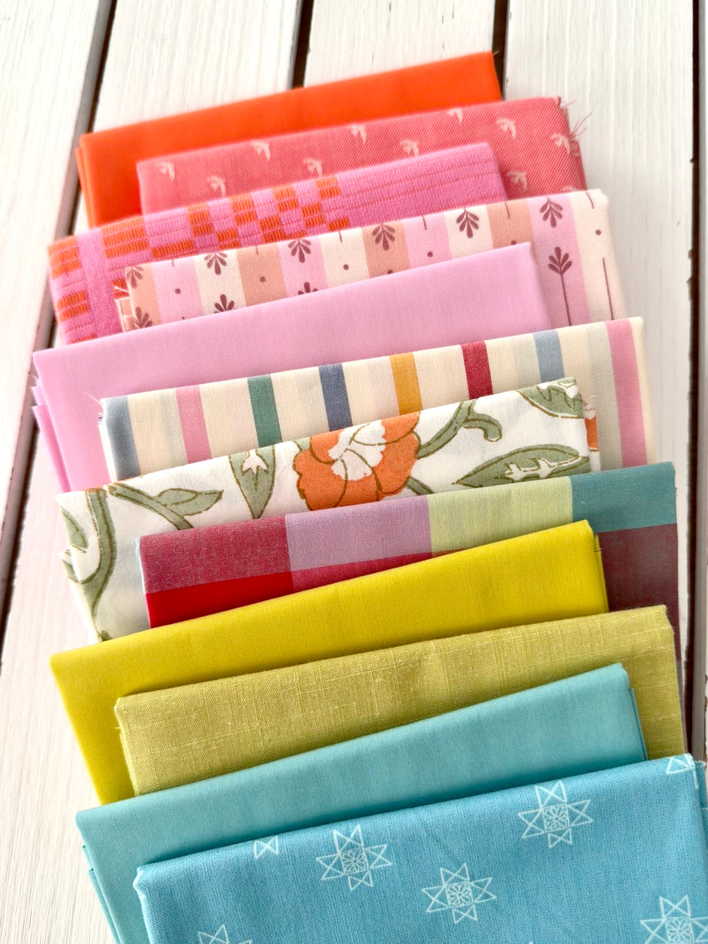 neatly folded set of fabrics stacked against a white wooden background. colors vary from bright orange-red, cream, chartreuse, and warm teals.