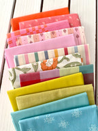 neatly folded set of fabrics stacked against a white wooden background. colors vary from bright orange-red, cream, chartreuse, and warm teals.