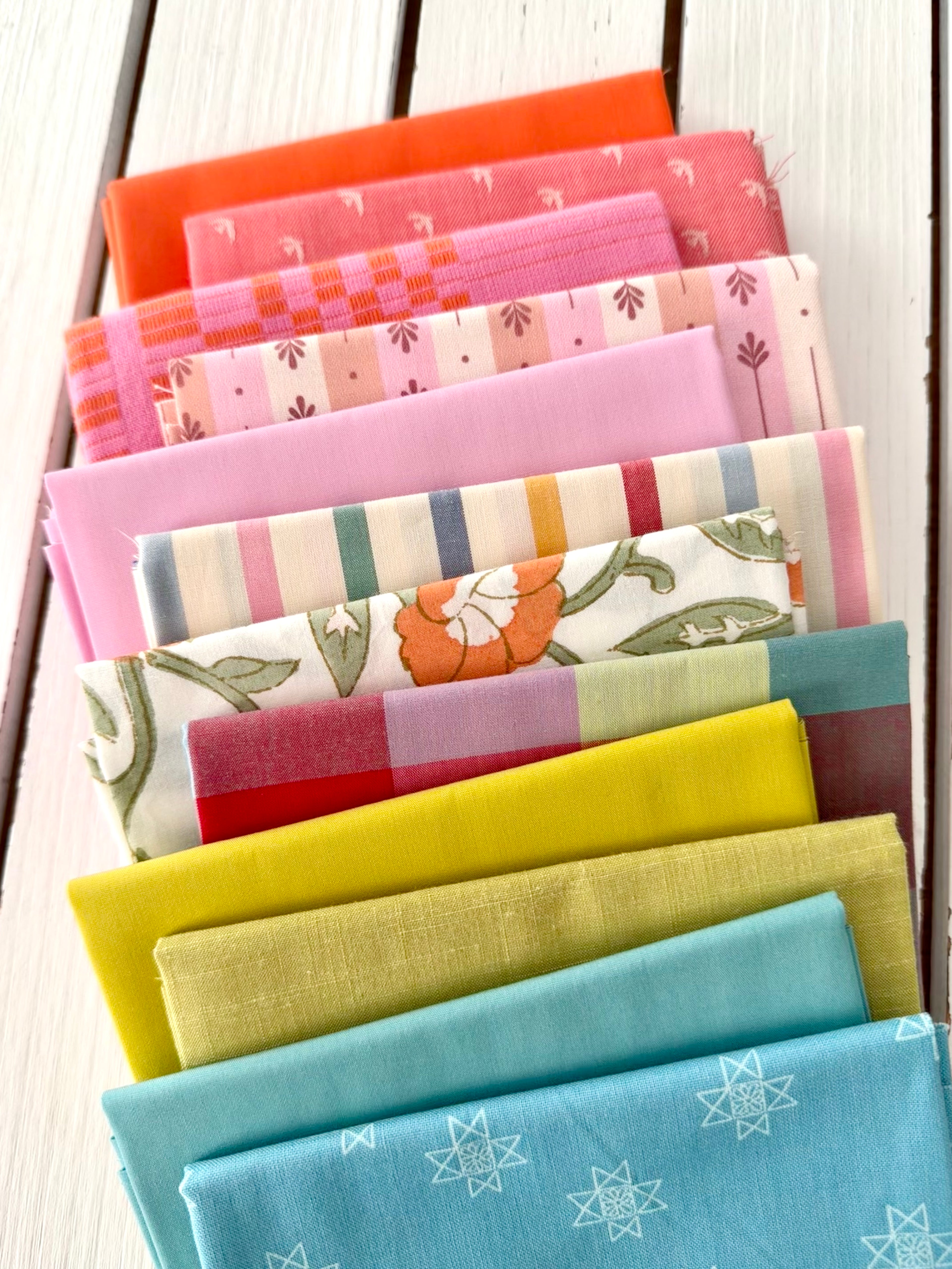 neatly folded set of fabrics stacked against a white wooden background. colors vary from bright orange-red, cream, chartreuse, and warm teals.