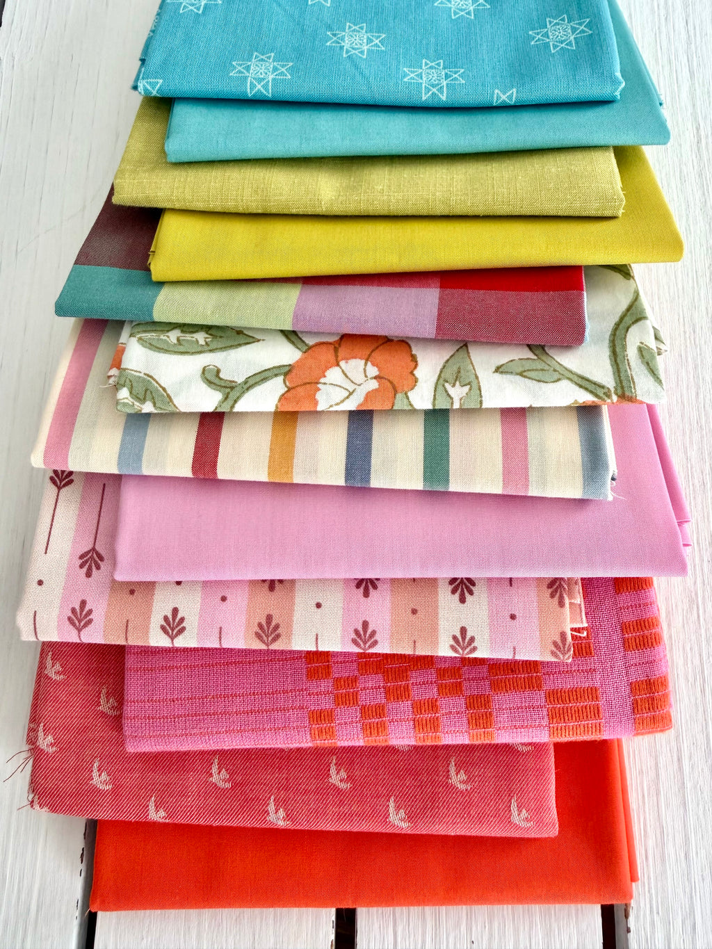 neatly folded set of fabrics stacked against a white wooden background. colors vary from bright orange-red, cream, chartreuse, and warm teals.