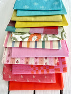 neatly folded set of fabrics stacked against a white wooden background. colors vary from bright orange-red, cream, chartreuse, and warm teals.
