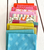 neatly folded set of fabrics stacked against a white wooden background. colors vary from bright orange-red, cream, chartreuse, and warm teals.
