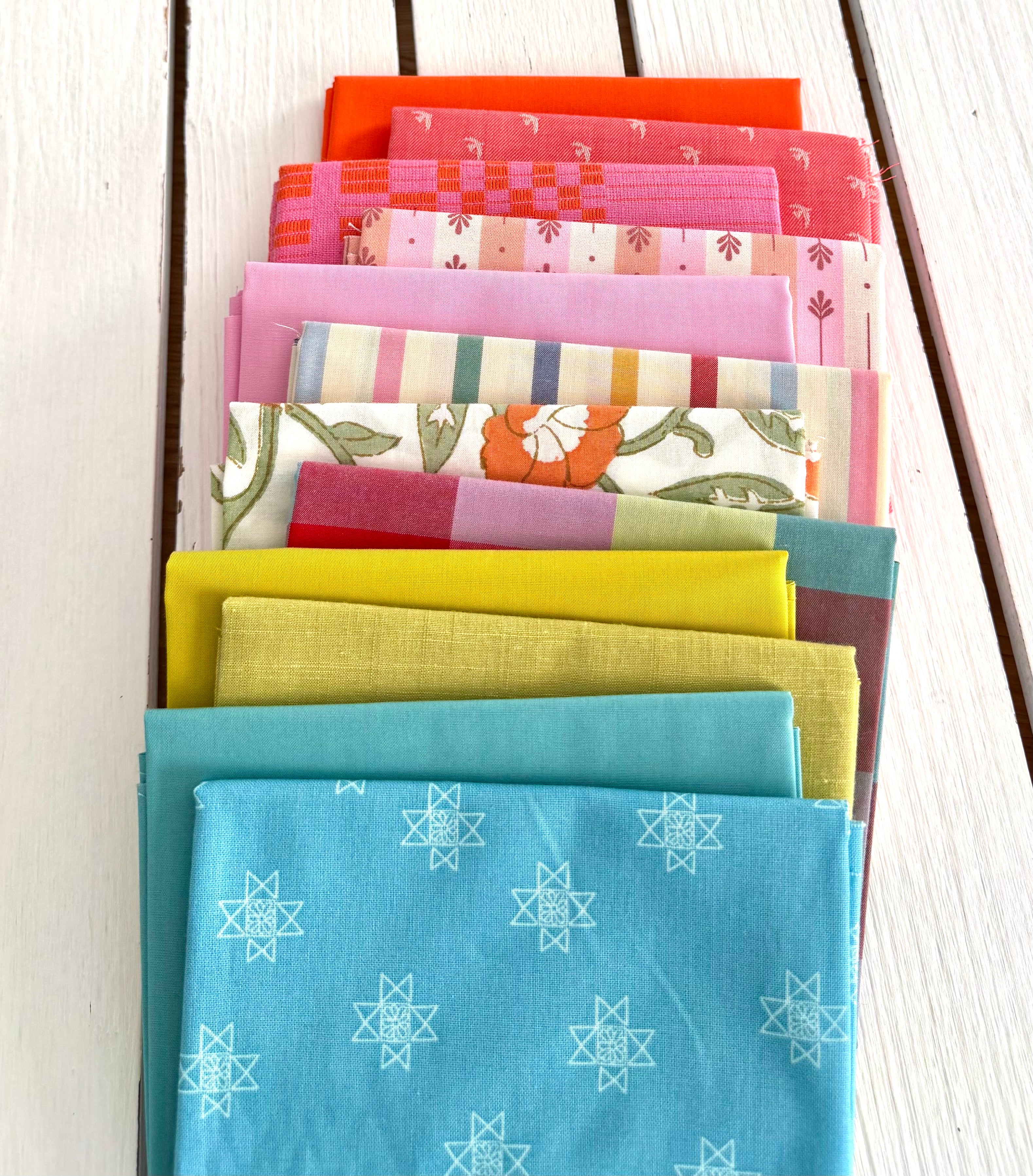 neatly folded set of fabrics stacked against a white wooden background. colors vary from bright orange-red, cream, chartreuse, and warm teals.