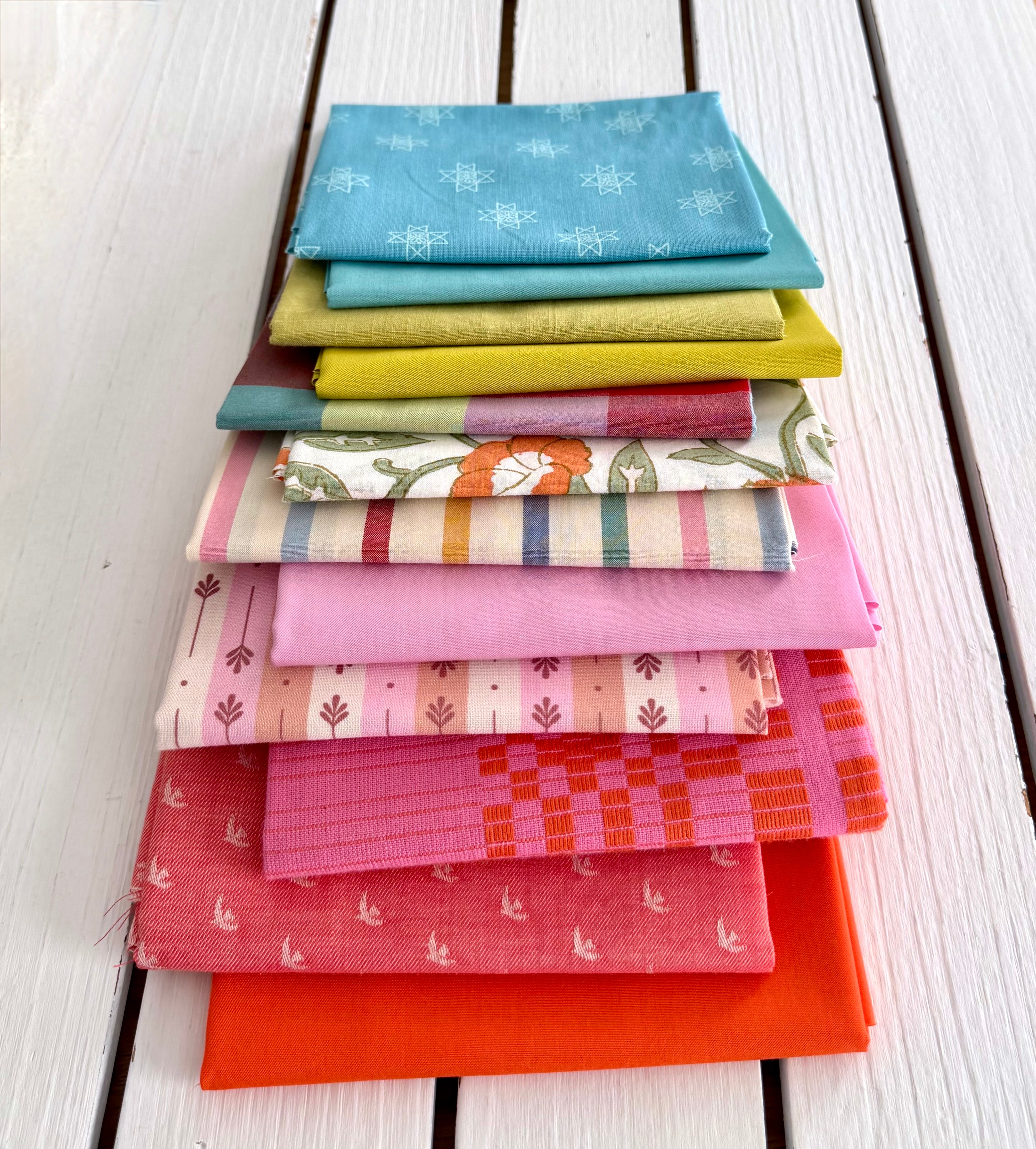 neatly folded set of fabrics stacked against a white wooden background. colors vary from bright orange-red, cream, chartreuse, and warm teals.