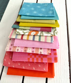 neatly folded set of fabrics stacked against a white wooden background. colors vary from bright orange-red, cream, chartreuse, and warm teals.