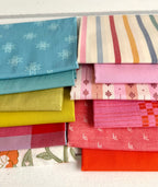 neatly folded set of fabrics stacked against a white wooden background. colors vary from bright orange-red, cream, chartreuse, and warm teals.